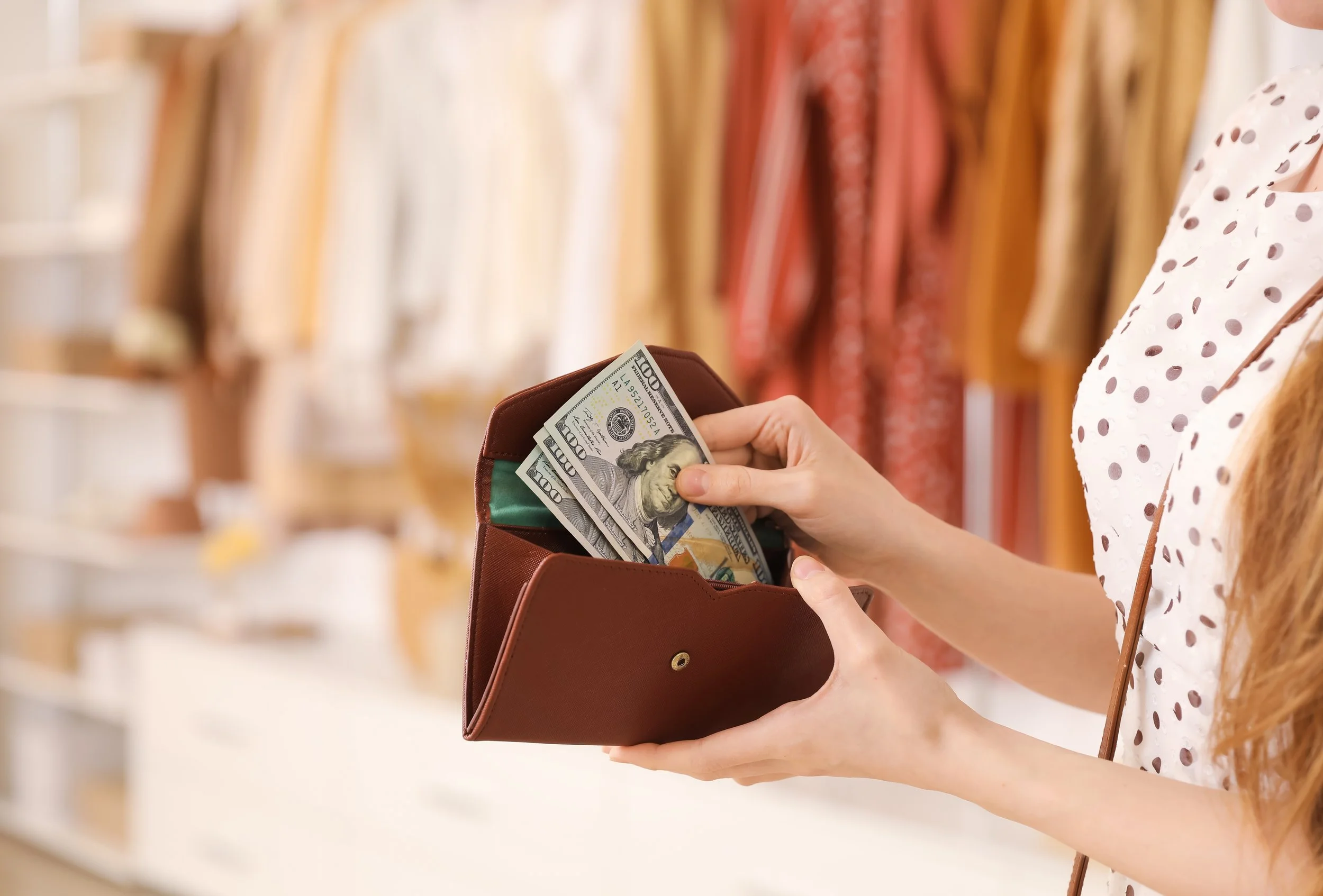 Woman holding cash in a wallet with soft-focus clothing rack background, symbolizing financial awareness, spending habits, and conscious budgeting