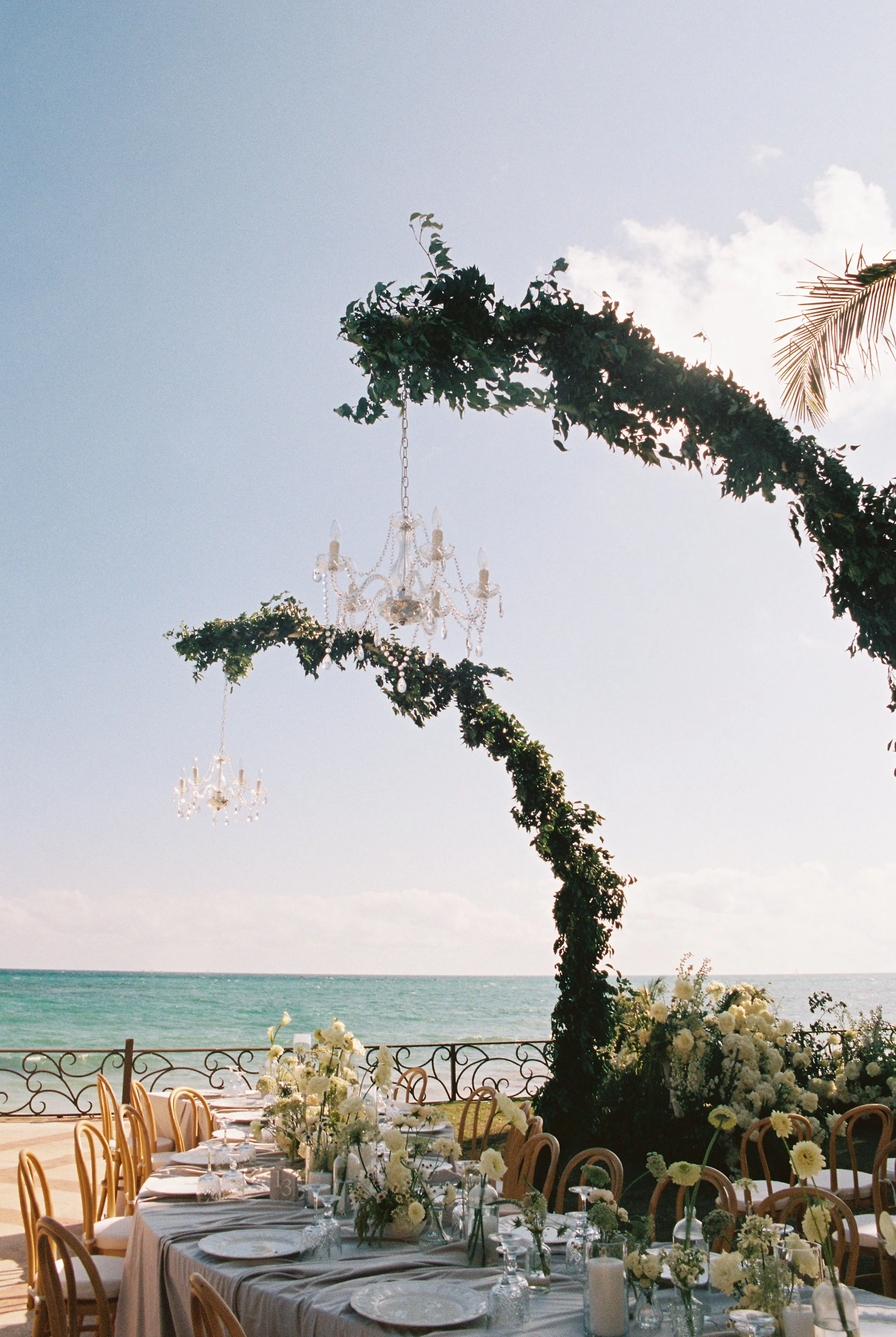 Ocean side reception serpentine tables with white florals and bud vases. Green floral arches with hanging crystal chandeliers