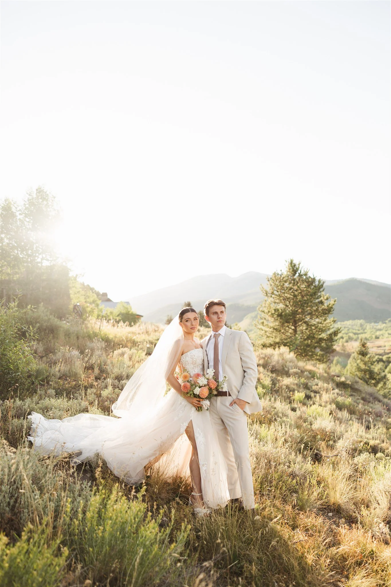 Styled bride and groom in a field mountainside with sun shining behind. Floral bouquet and lace detailed dress