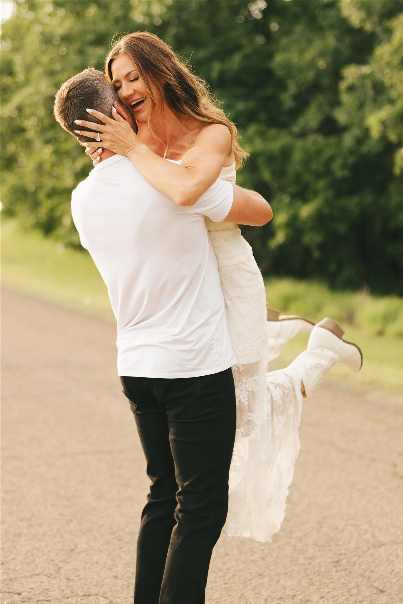 couples engagement photos outside with greenery and roadside in the background, woman wearing white dress and cowgirl boots