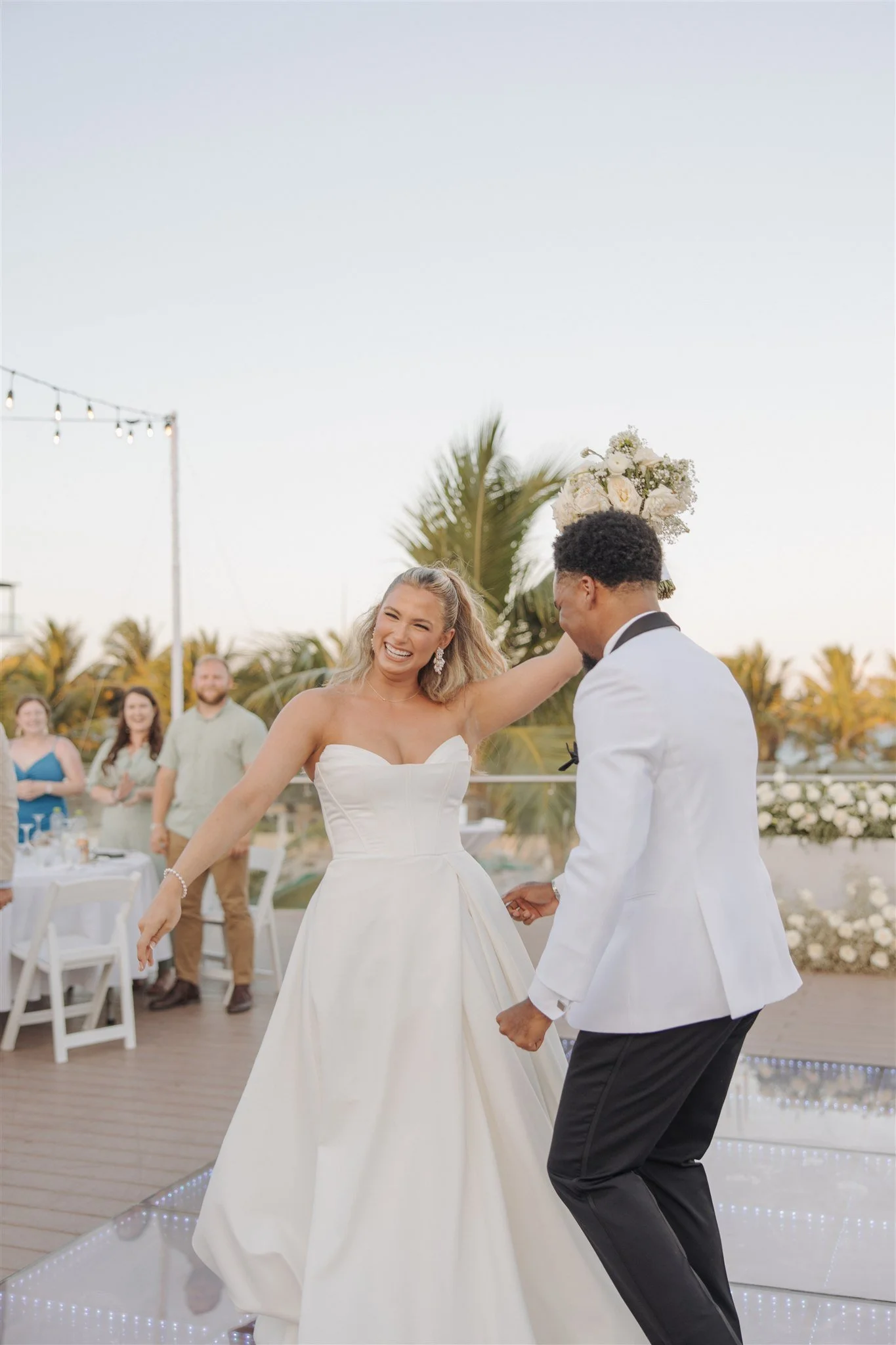 Destination bride and groom having a fun moment during their first dance and celebrating with their guests. Bride holding a white floral bouquet and a-line style dress, Groom in white suit jacket and black pants.