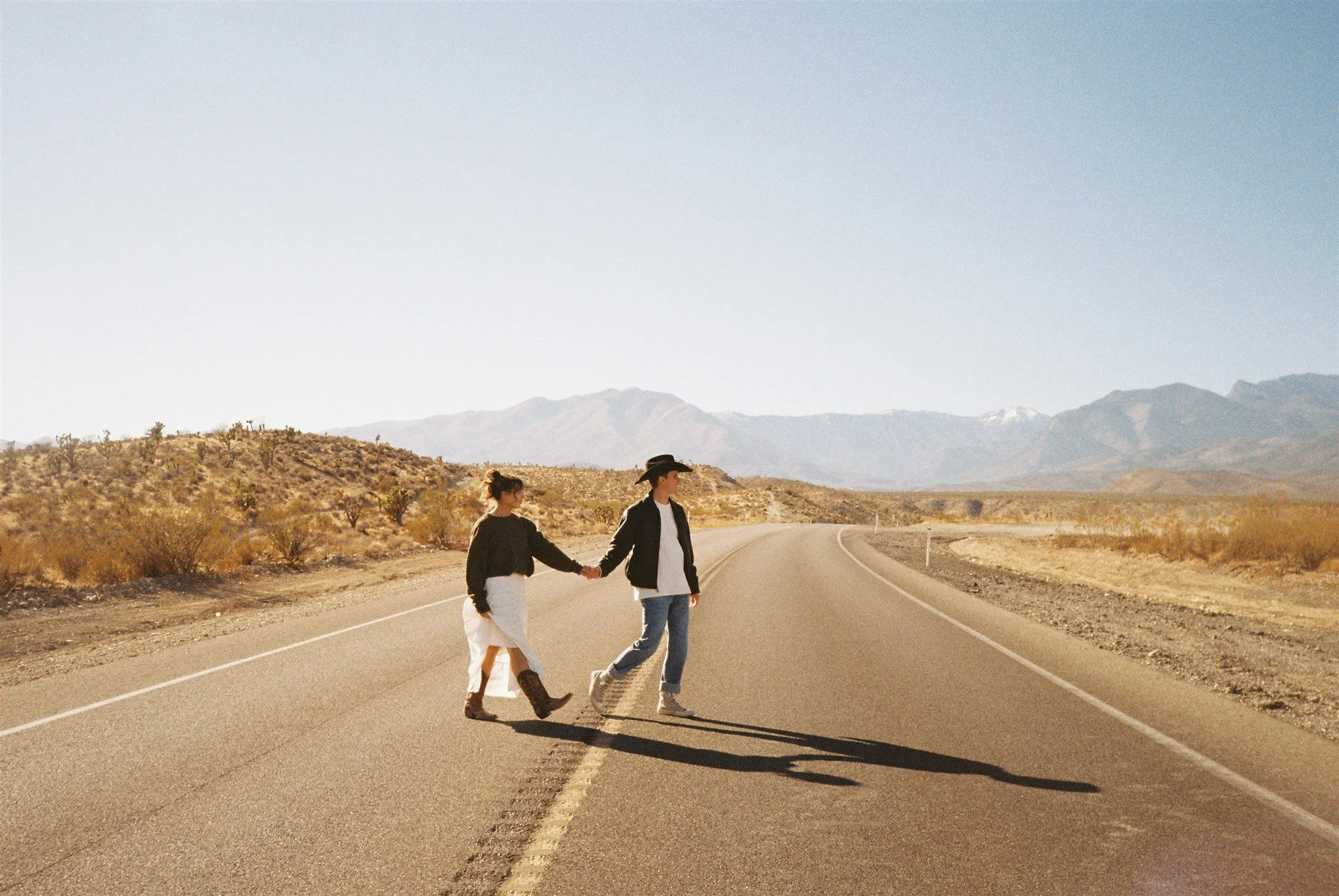 Couples photo with a man and woman grasping hands as they cross a dessert road. cowgirl boots, rustic vibe, and mountain views