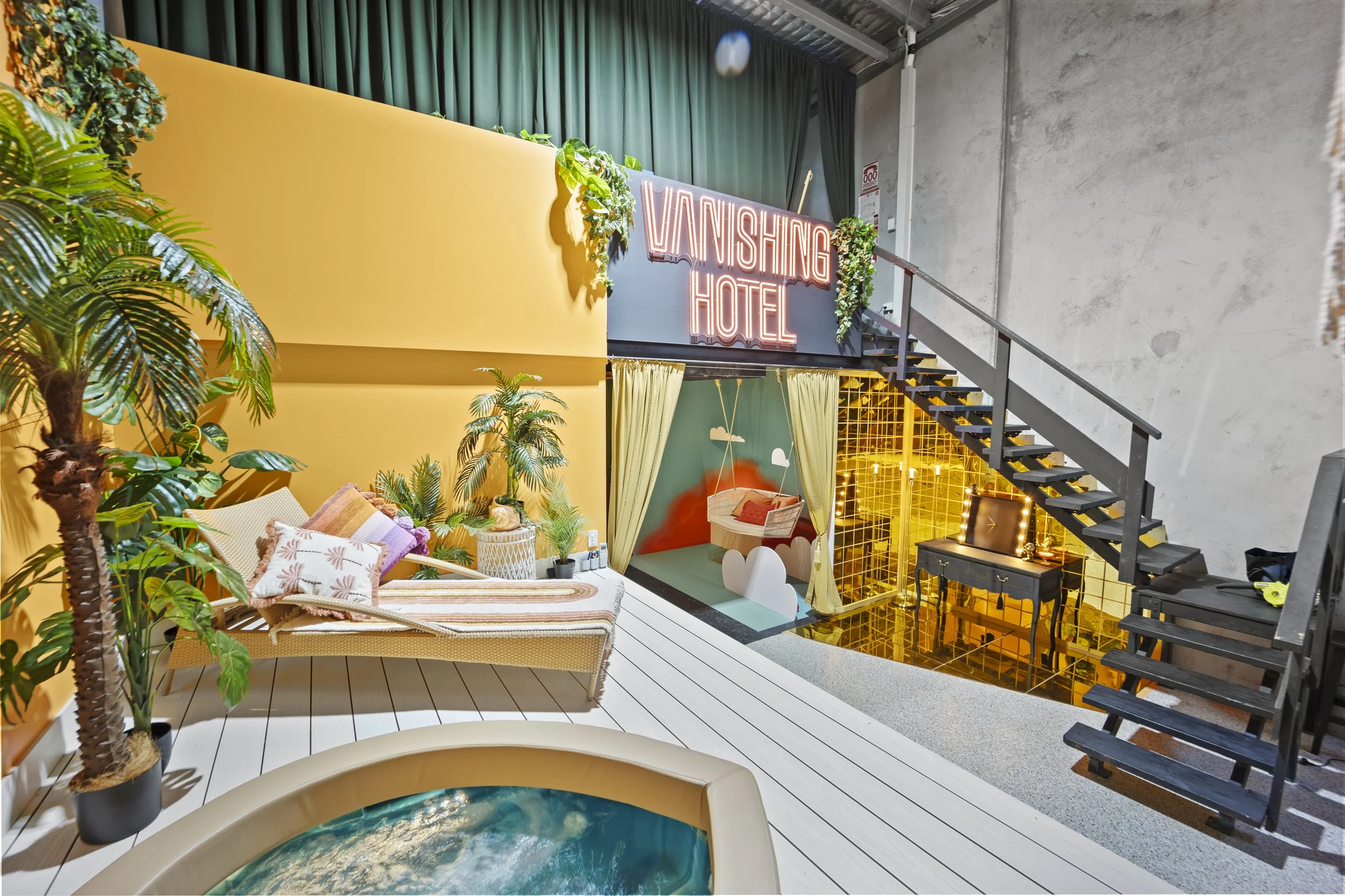 Interior of a studio labeled "Vanishing Hotel" with tropical decor, lounge chair with cushions, small pool, swing bed, stairs, and a mirrored makeup area.