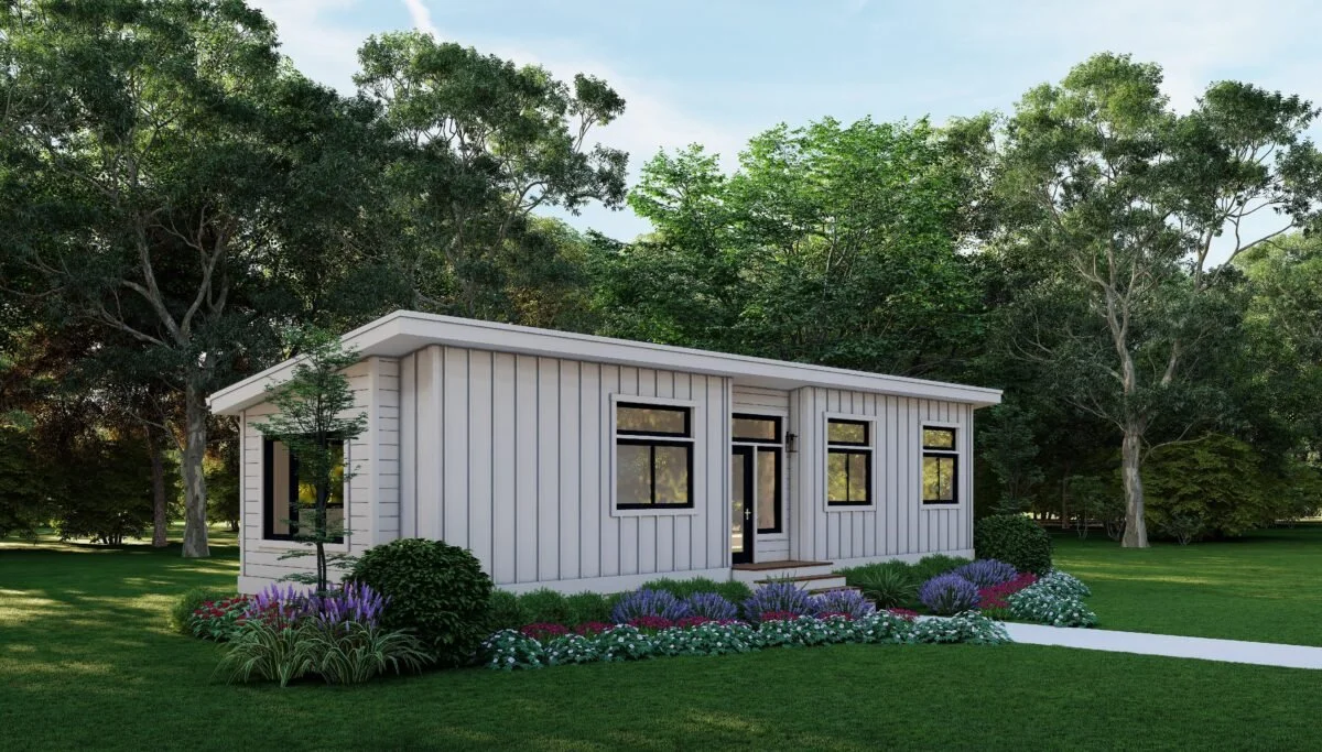 A small, white, modern house with vertical siding, black framed windows, and a glass front door, surrounded by landscaped flower beds and large trees in the background.