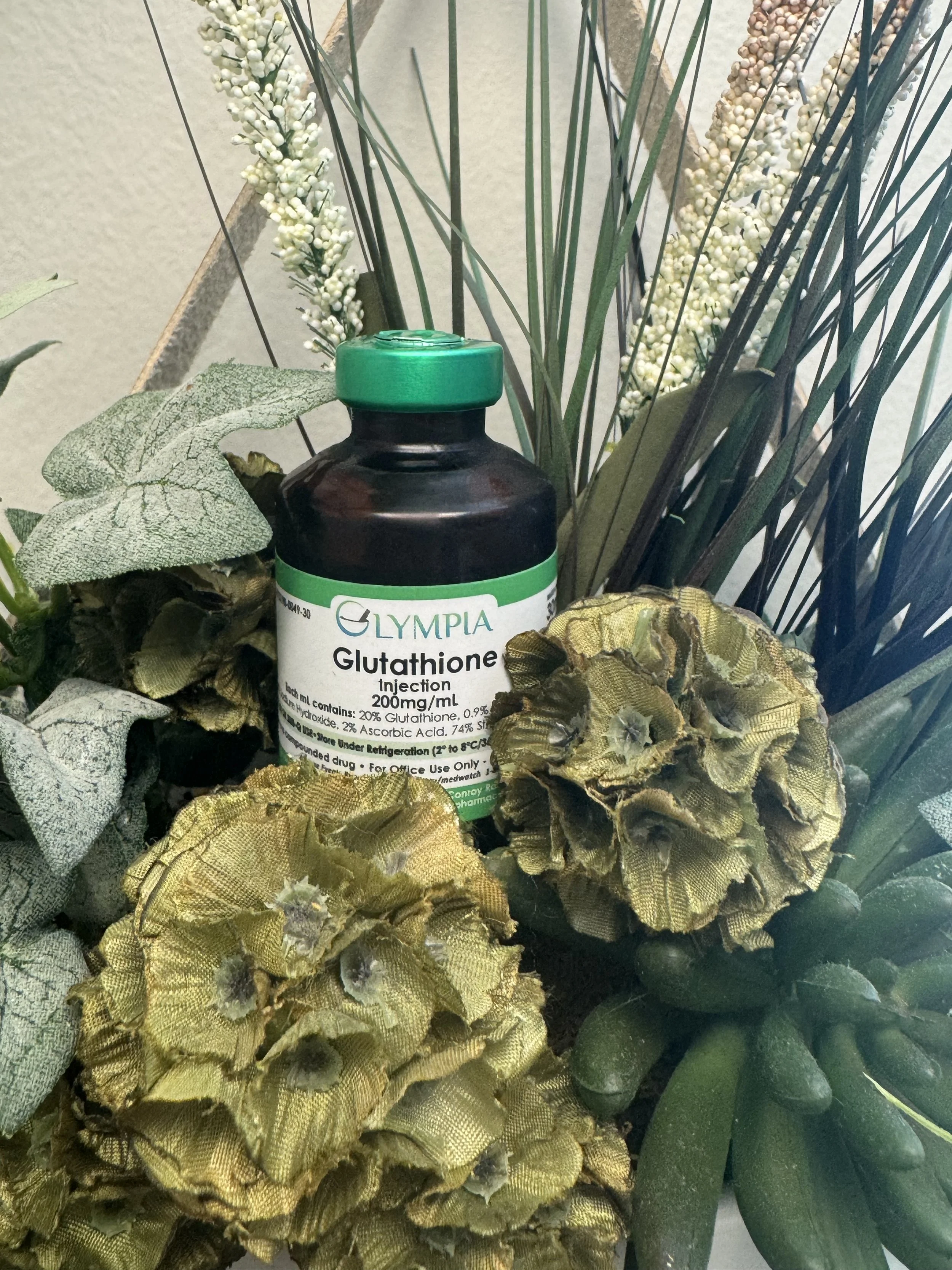 A black bottle labeled 'Glutathione Injection 200mg/ml' surrounded by artificial leaves and flowers in a decorative arrangement.