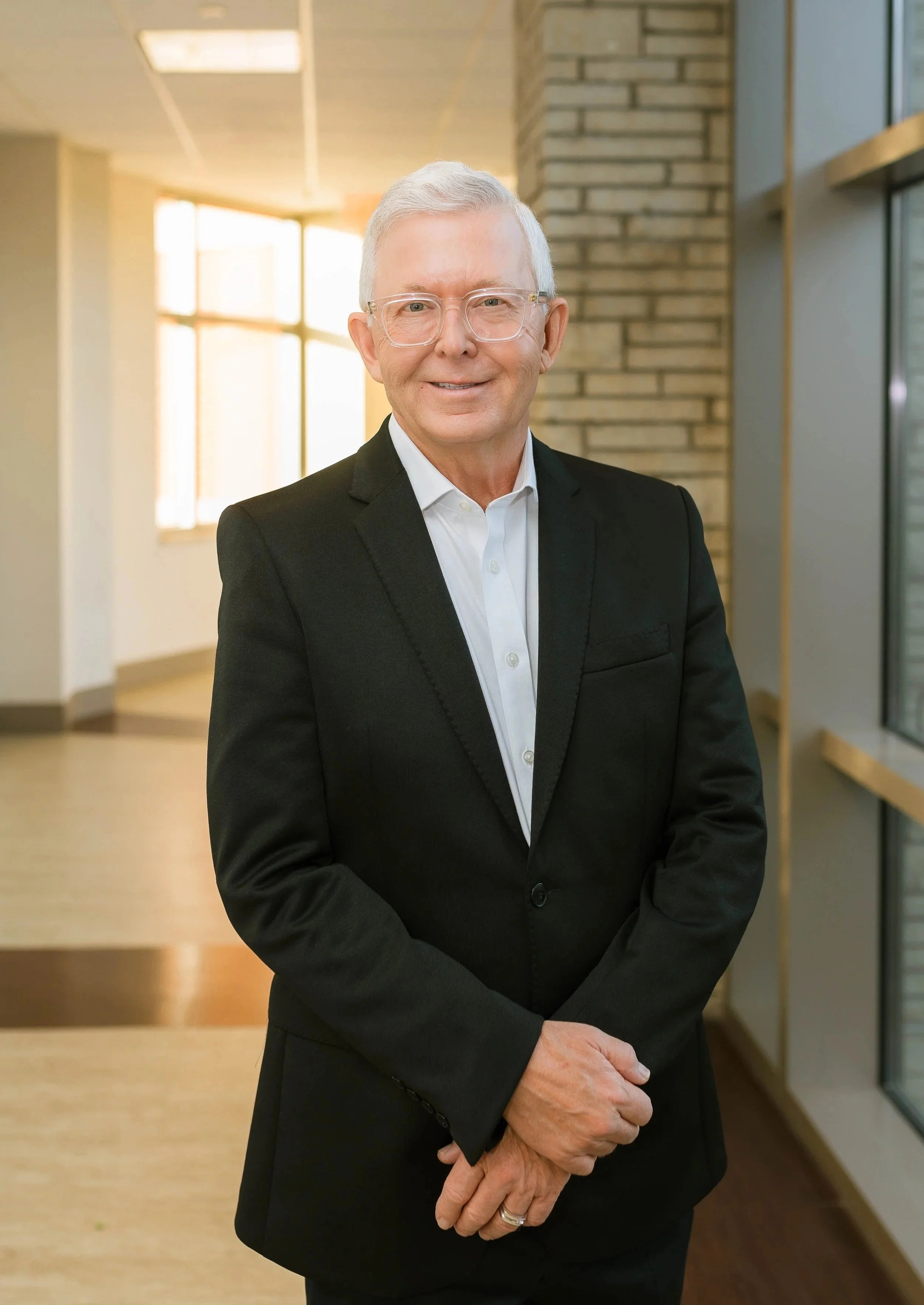 Dr Carter Abbott MD wearing glasses, a white shirt, and a black suit stands in a modern office hallway with large windows, brick wall accents, and natural light.