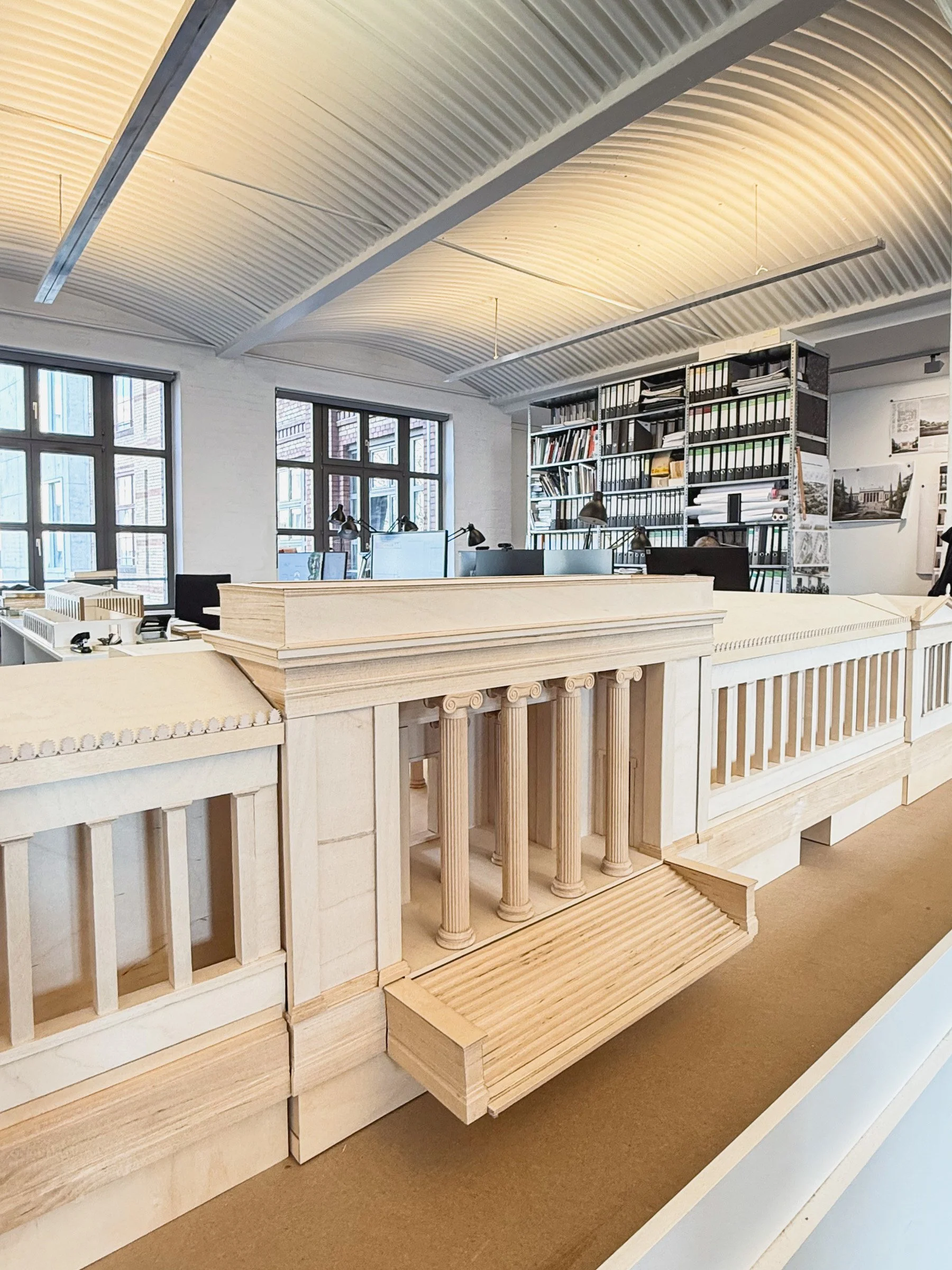 A wooden architectural model of a building interior featuring columns, a staircase, and shelving units with desks and computers near windows.