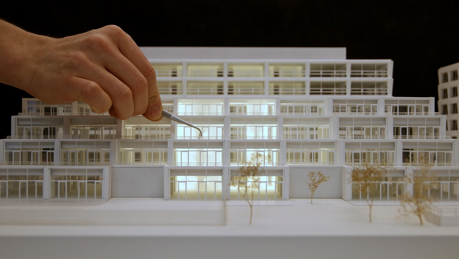 A person using a fine tool to work on a detailed architectural model of a multi-level building, with trees in front.