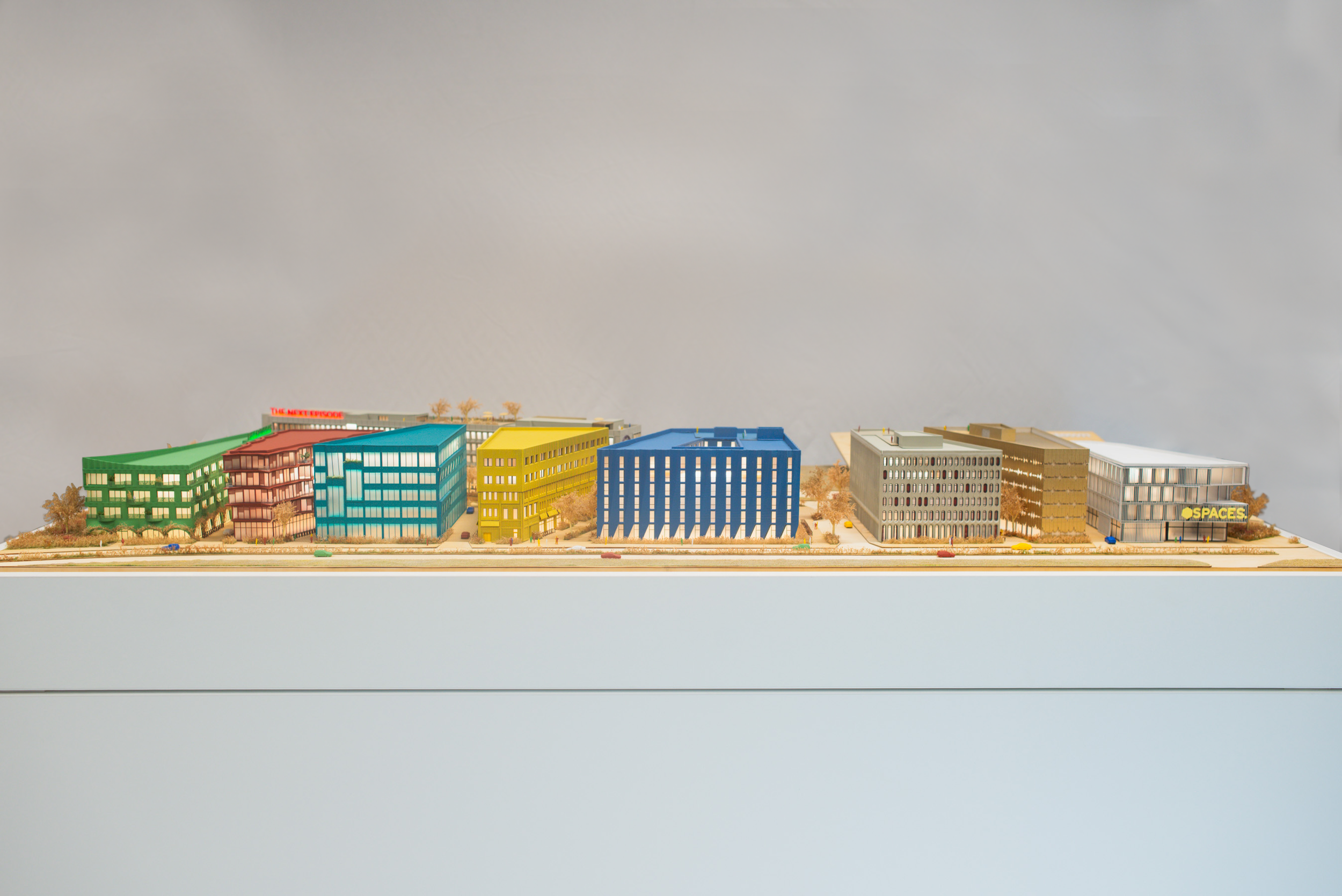 A detailed miniature model of a city street with colorful buildings, trees, and a sidewalk, set against a plain, light-colored background.