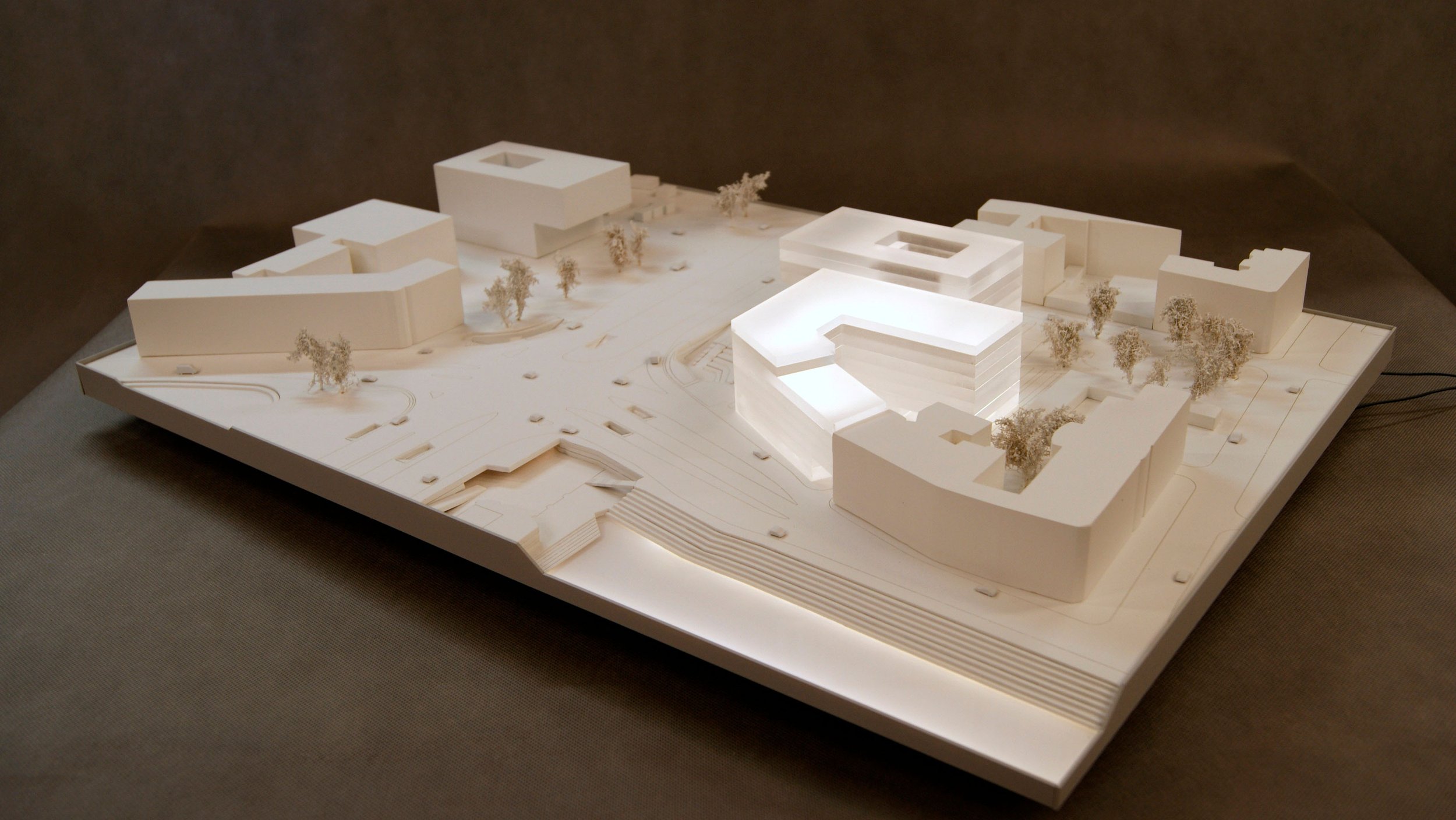A detailed architectural scale model of a modern building complex, illuminated with built-in lighting, with trees around it and roads surrounding the site.