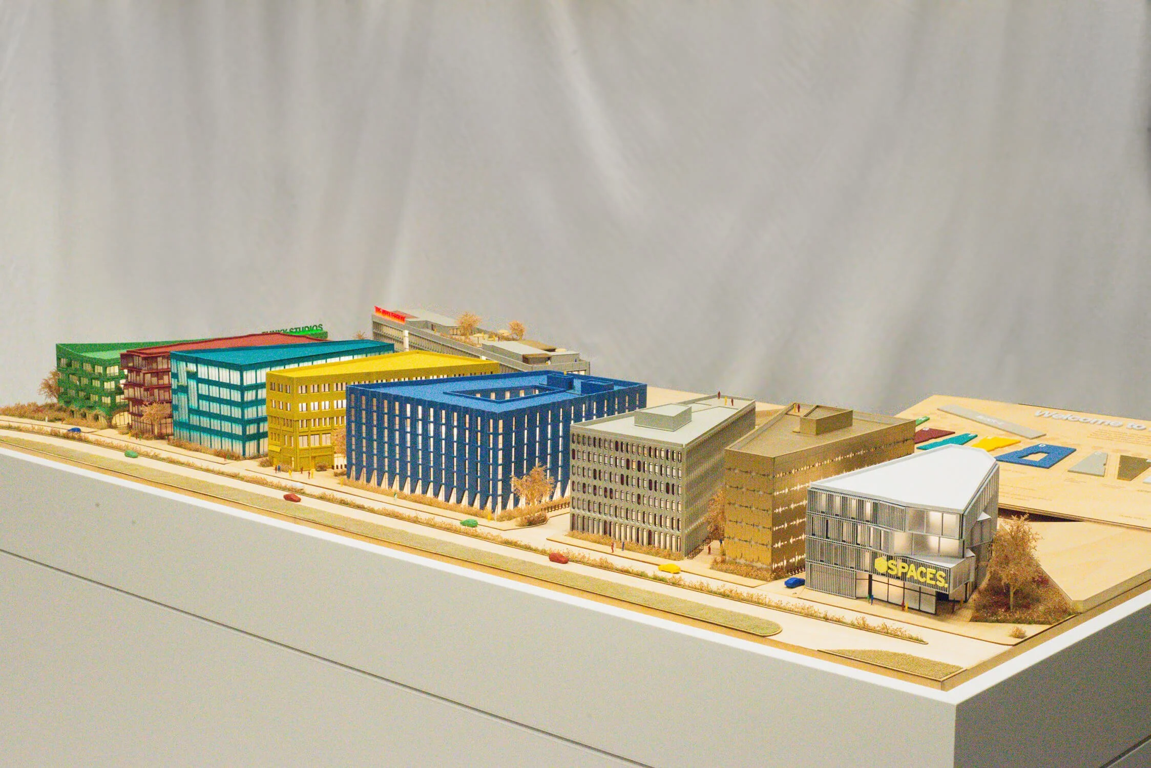 Architectural model of a modern urban development with colorful buildings, trees, cars, and minor figure models on a white display table.