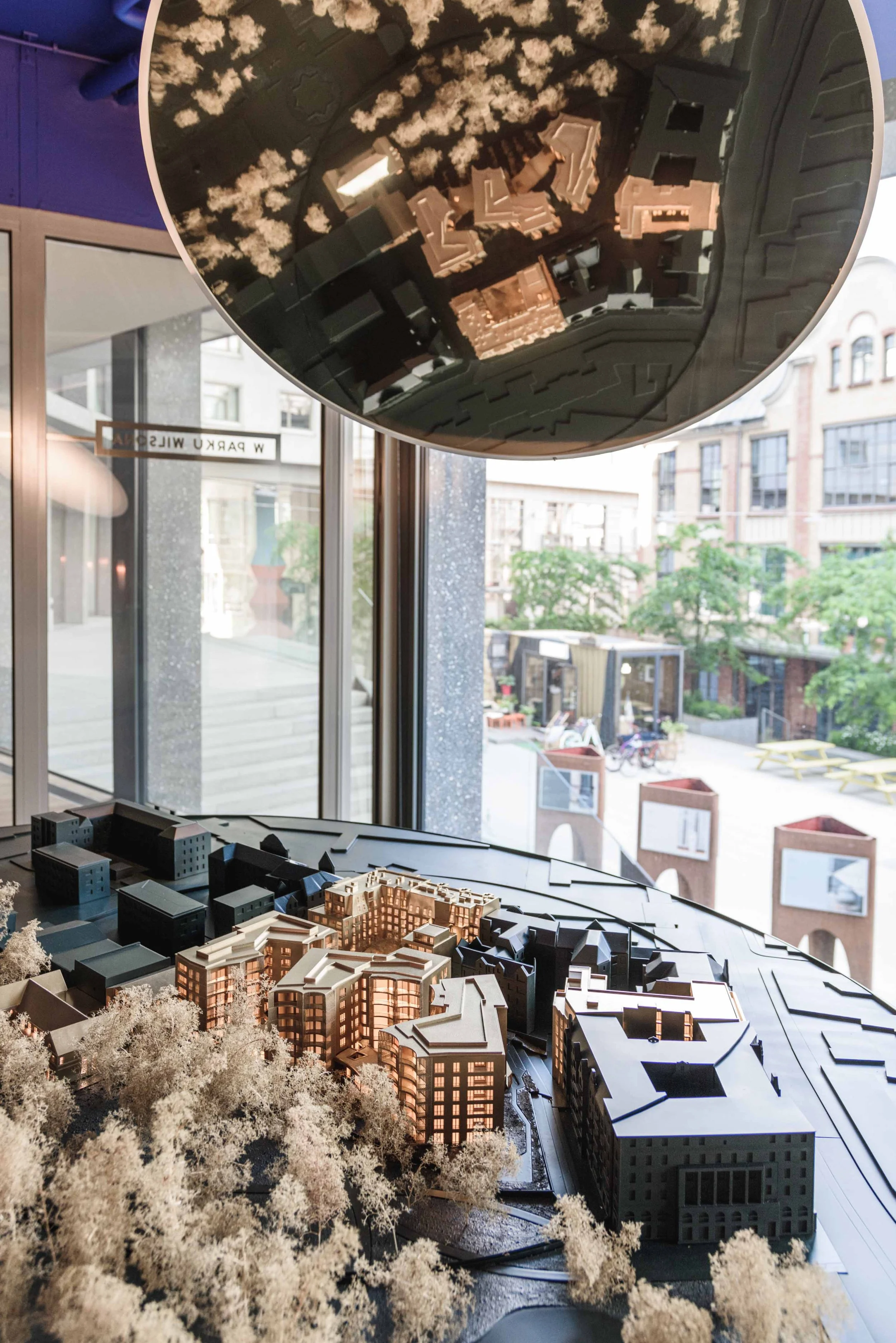 A detailed architectural scale model of a city block, featuring multiple buildings with varying architectural designs, located indoors near large windows with a cityscape view outside.