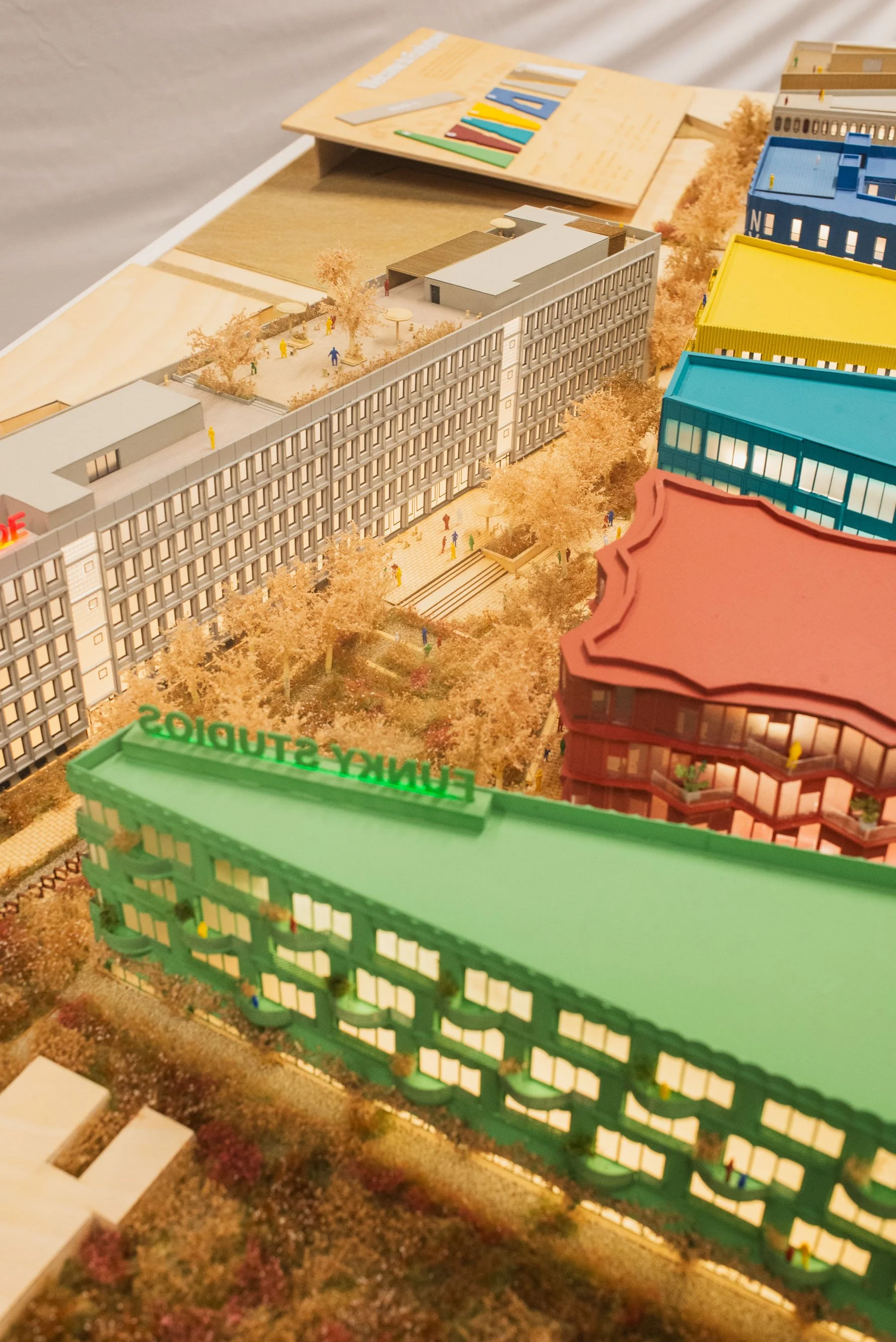 A detailed architectural model of a multi-story urban development with colorful buildings, trees, and pathways, including miniature human figures and outdoor seating areas.