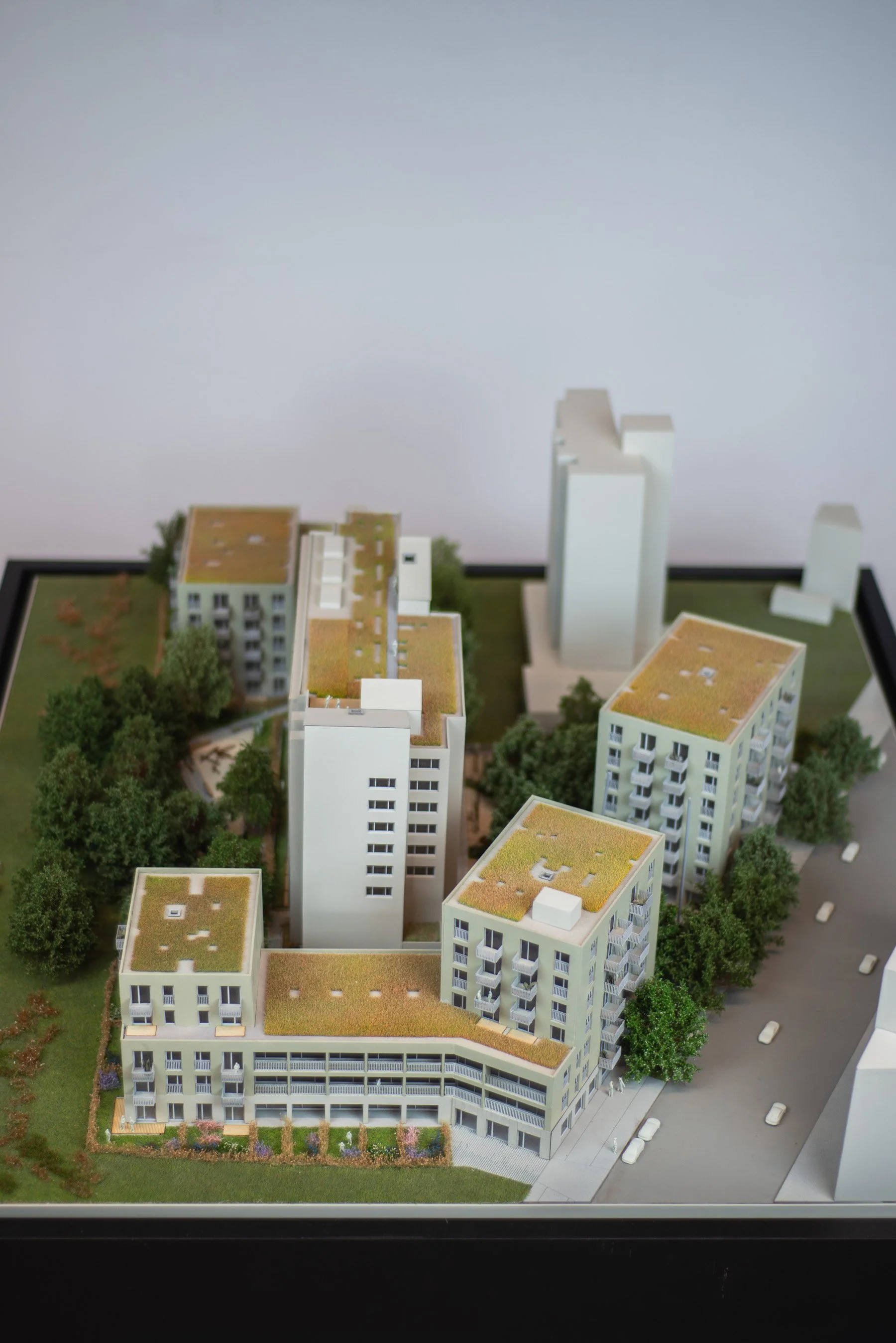 A detailed architectural model of a modern residential complex with multiple white buildings and green spaces, trees, and a surrounding street with parked cars.