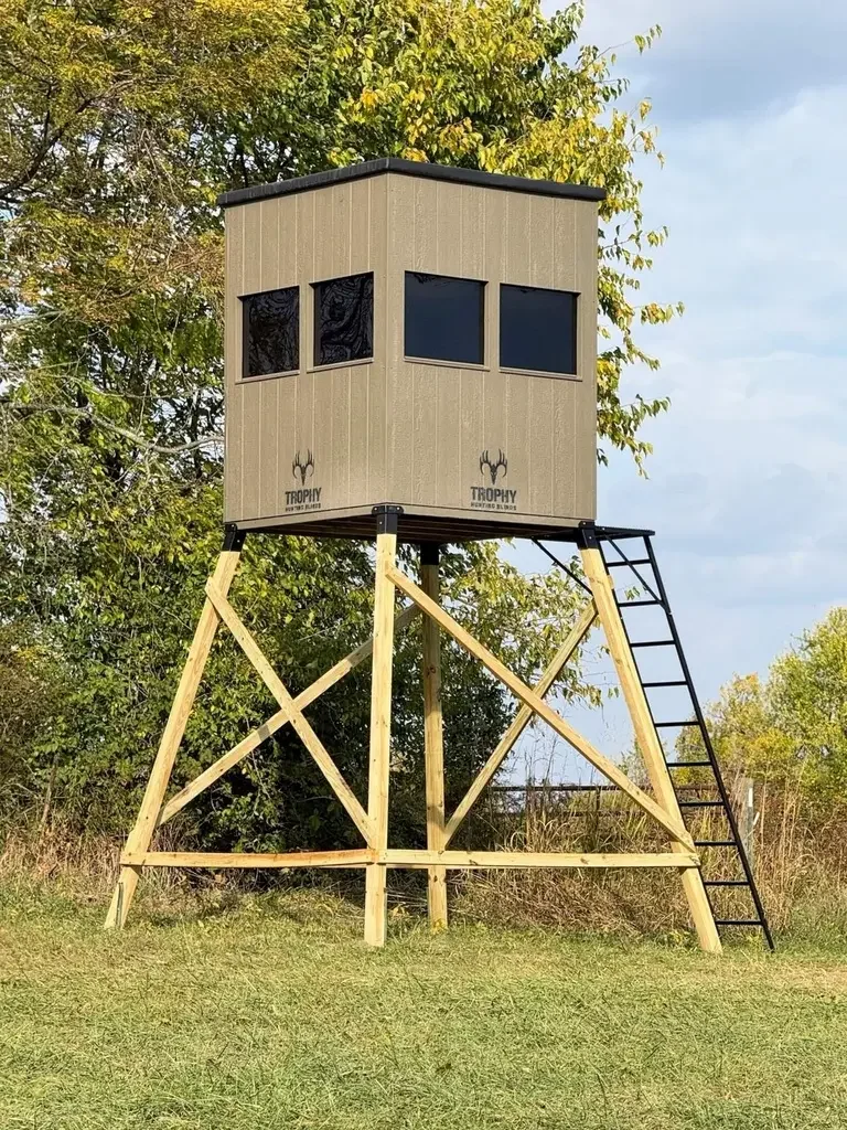 A hunting blind elevated on a wooden platform with a ladder, surrounded by trees and grass.