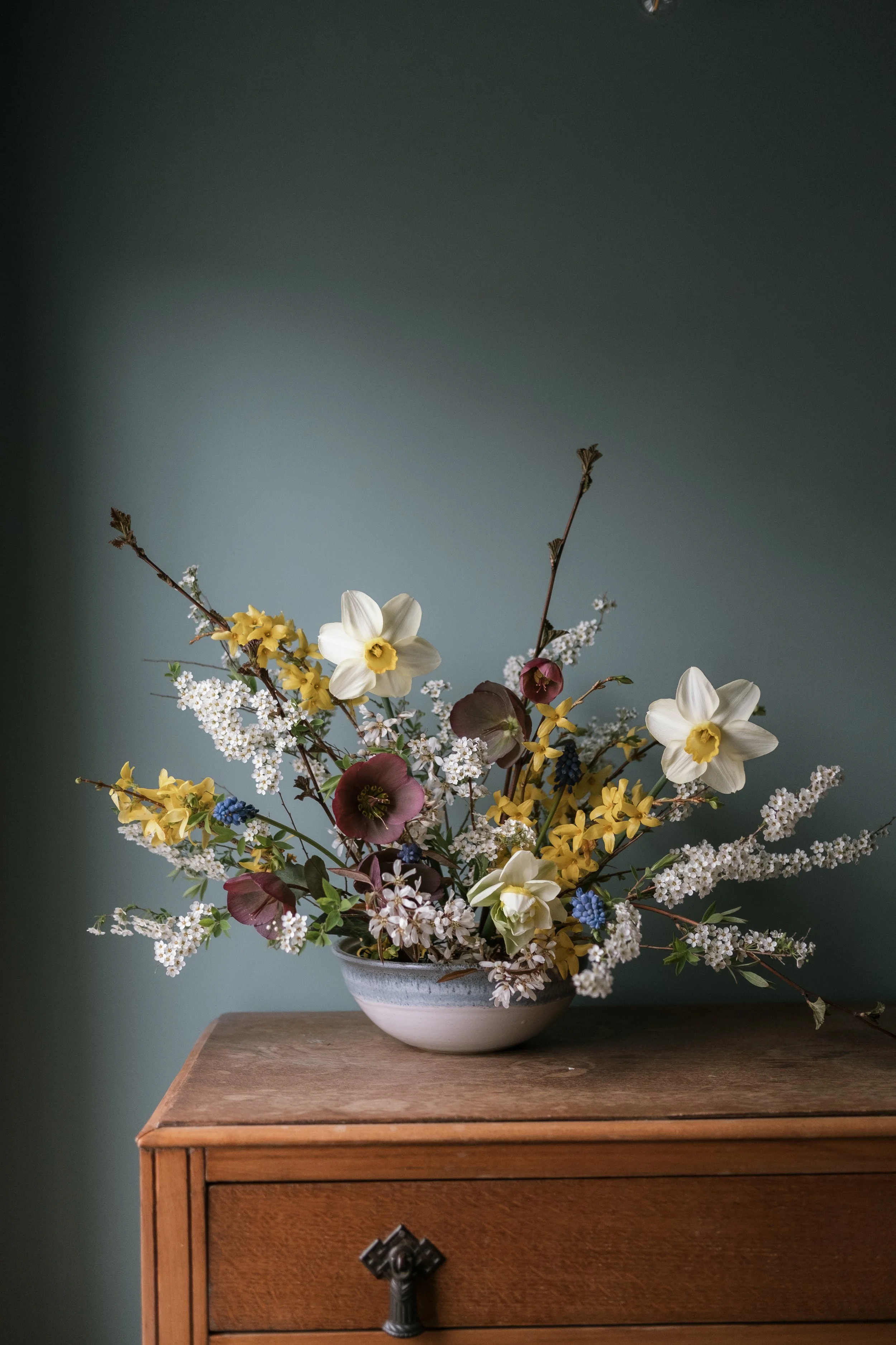 A floral arrangement in a white bowl placed on a wooden table, with a dark green wall in the background.