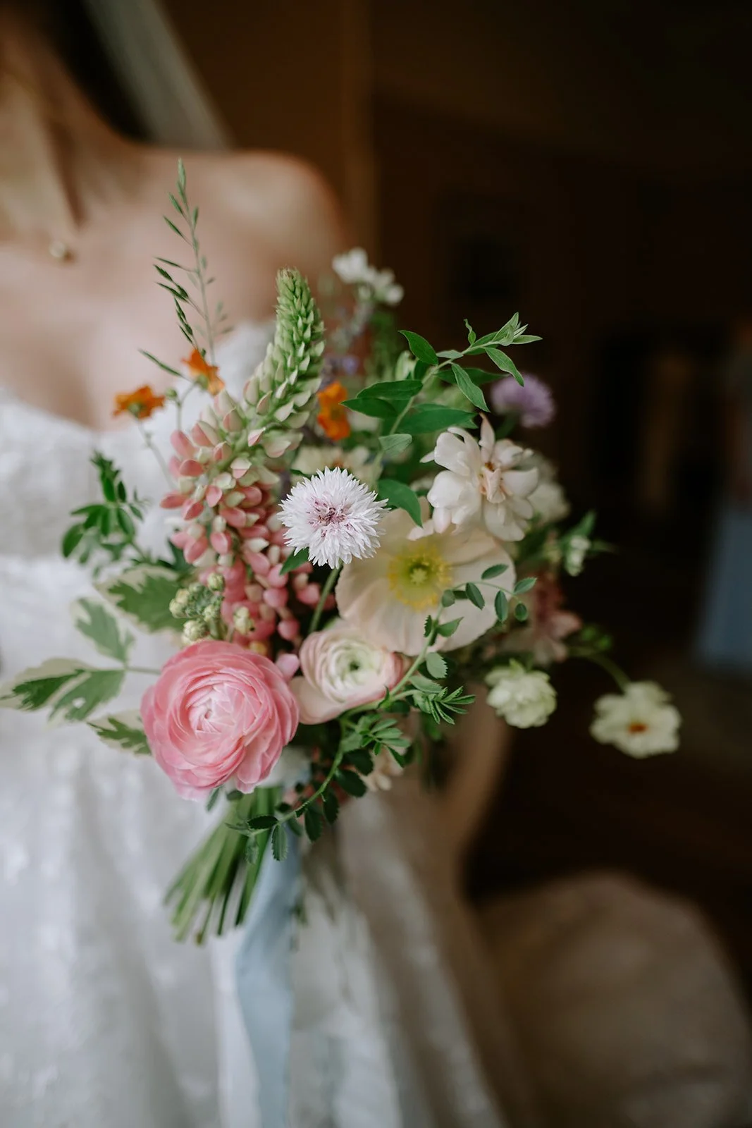 Close-up of a woman holding a bouquet of mixed pink, white, and orange flowers with green leaves, dressed in a white strapless garment.