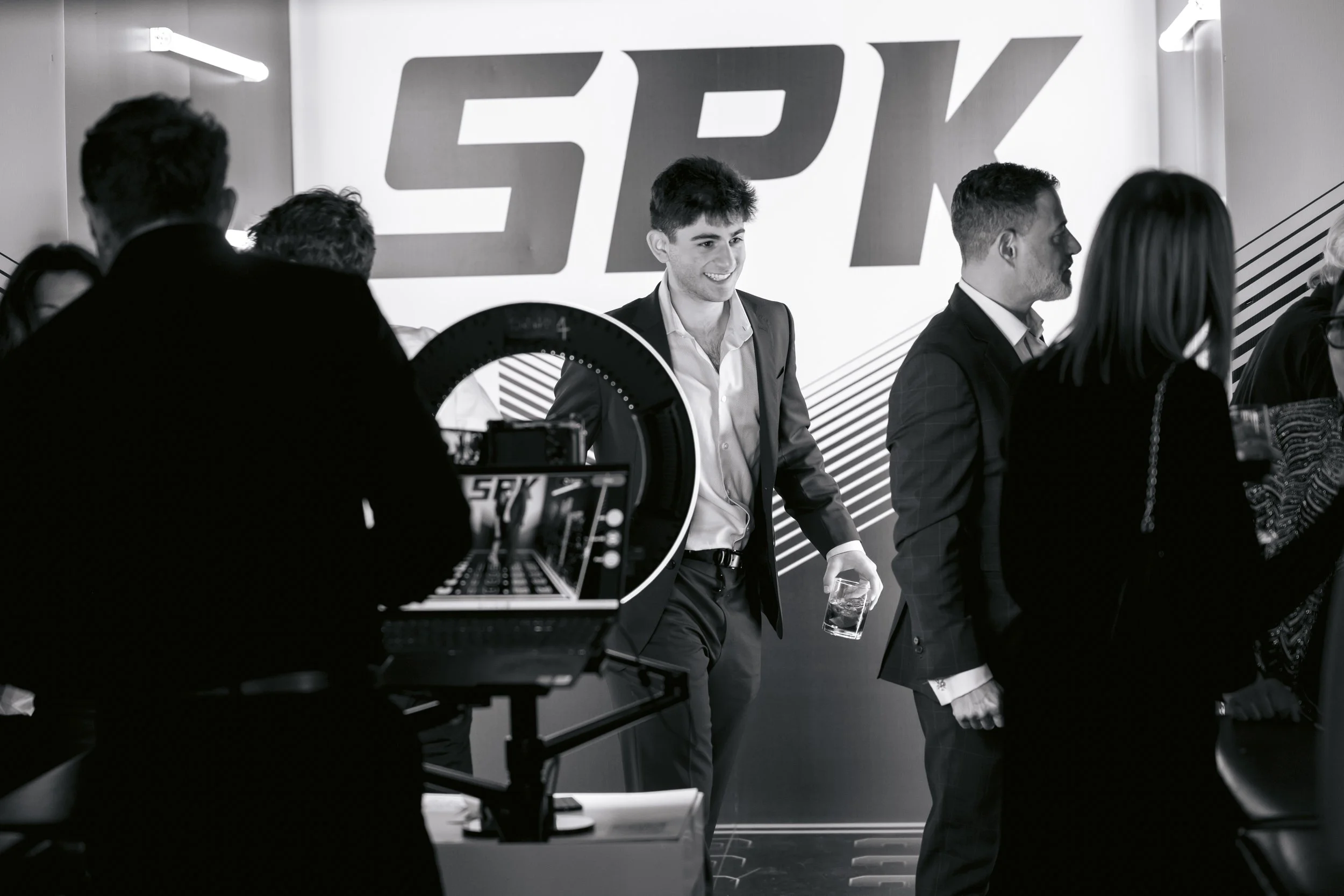 Men and women at a corporate event or conference, with a large branded backdrop reading 'FPK', some engaging in conversation, and a person operating a camera and laptop in the foreground.