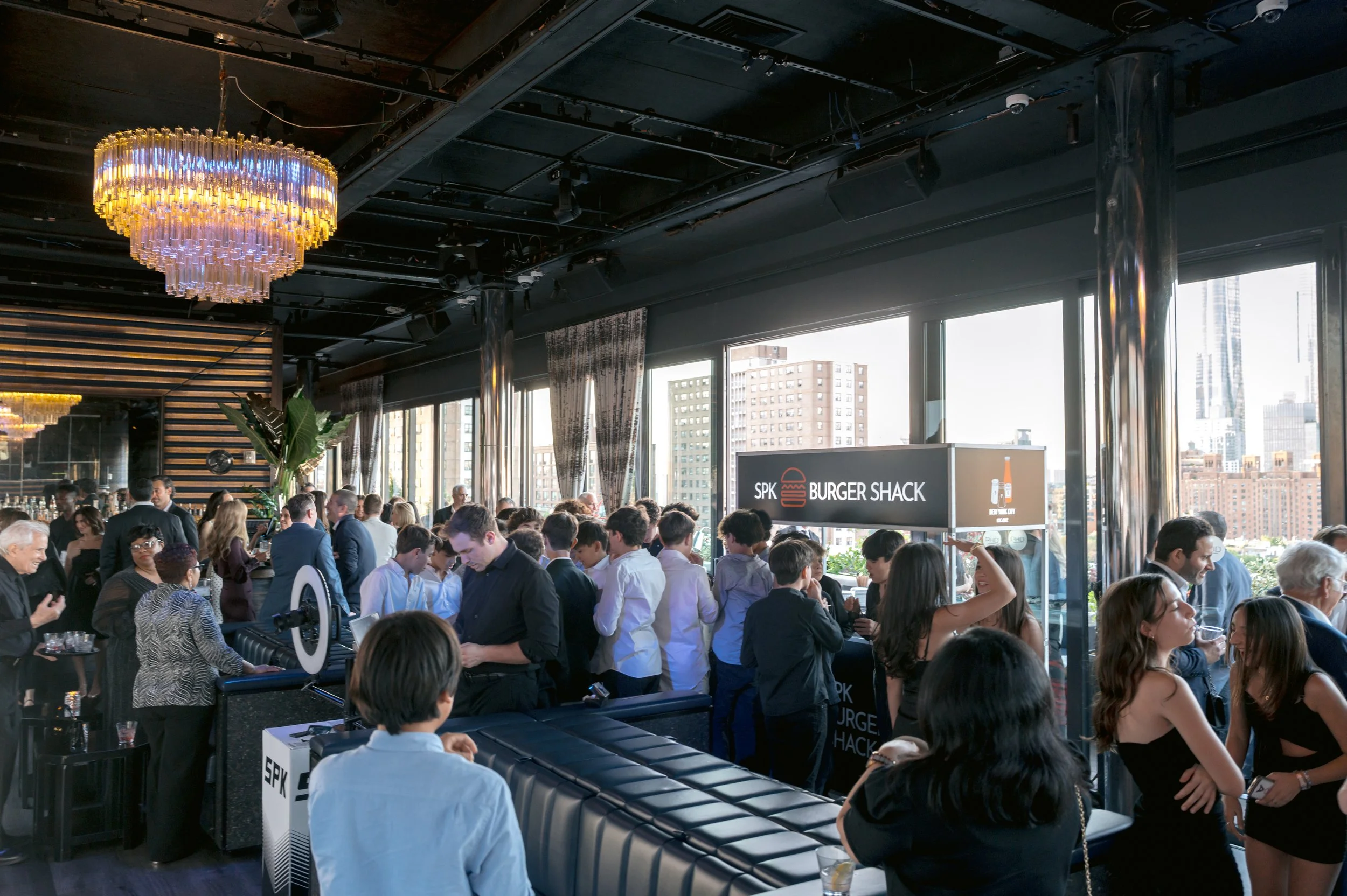 Crowd of people inside a modern restaurant with large windows, city skyline view, dim lighting, chandelier, and a Burger Shack sign.