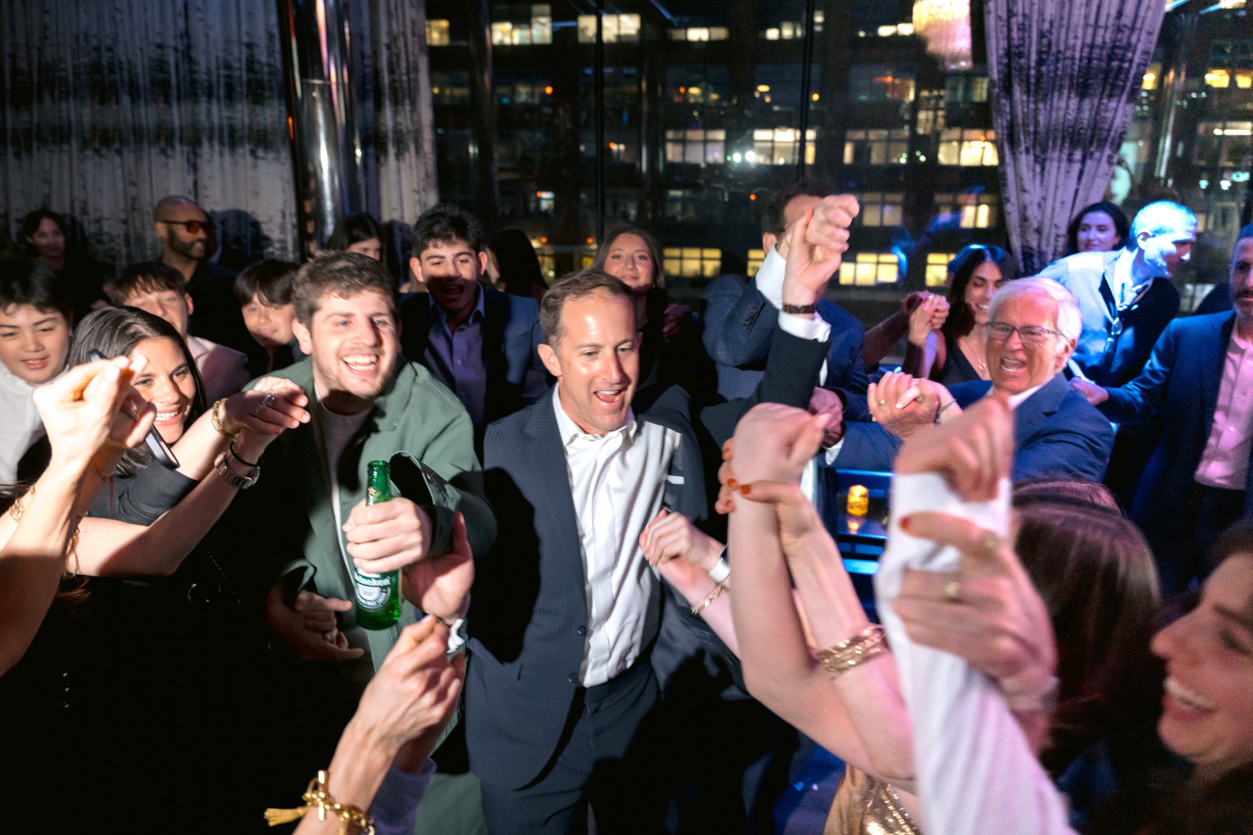 Group of people dancing and celebrating at a party or event, indoors at night with city lights visible through large windows in the background.