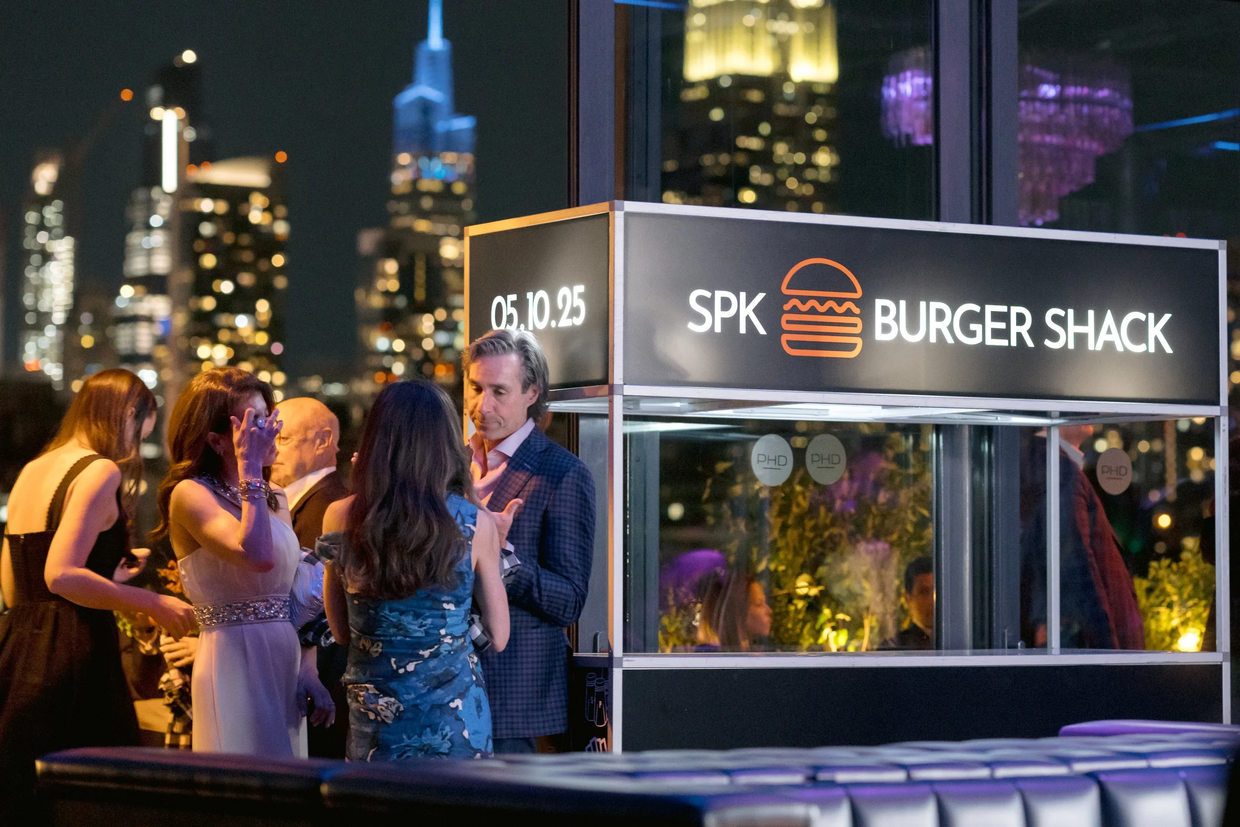 People gathered around a Burger Shack food stand at night with city skyline in the background.