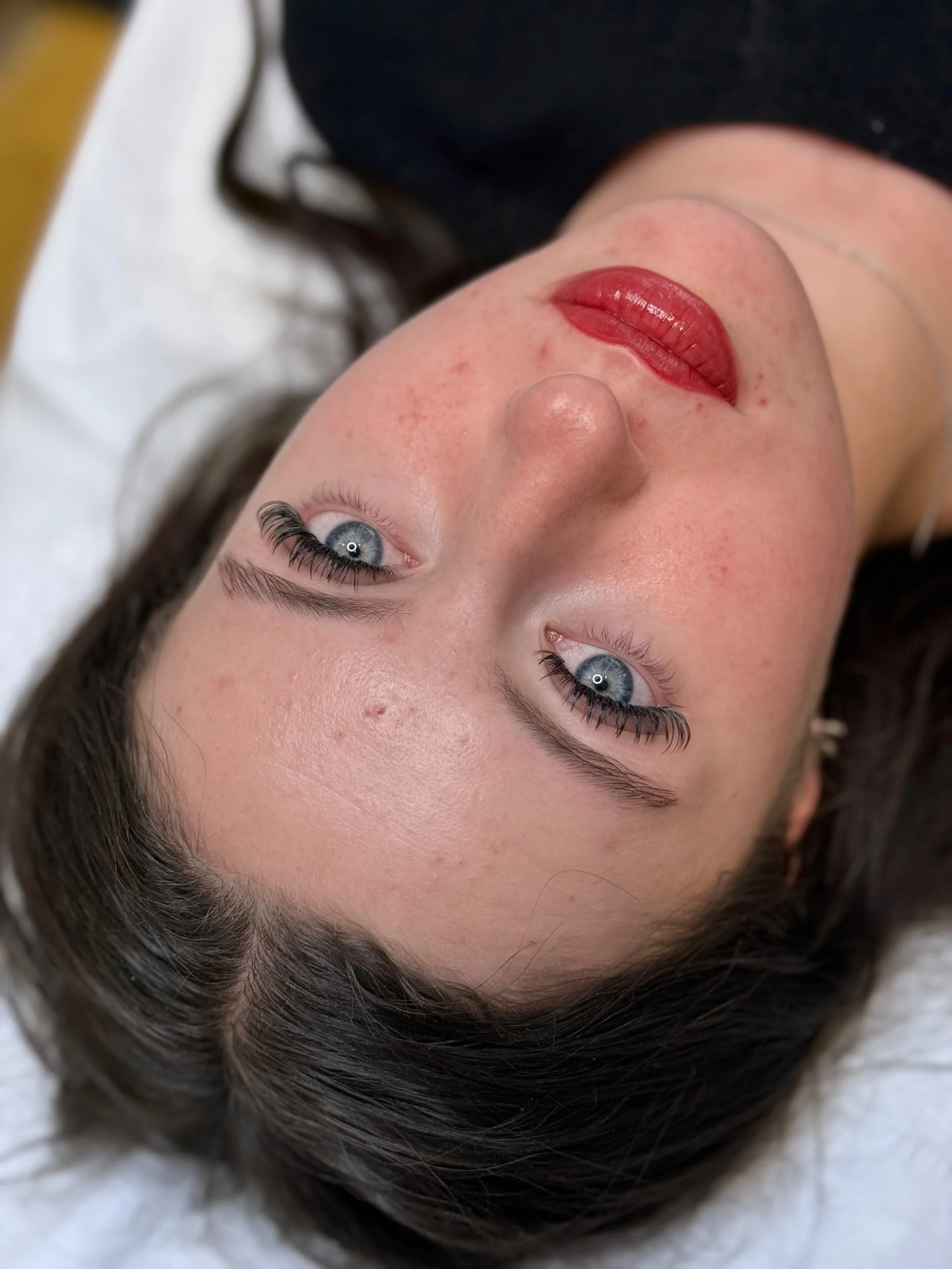 Lip Blush Cambridge — A close-up of a woman's face lying on a white surface, with blue eyes, dark wavy hair, and red lipstick  — Cambridge Brow Sanctuary