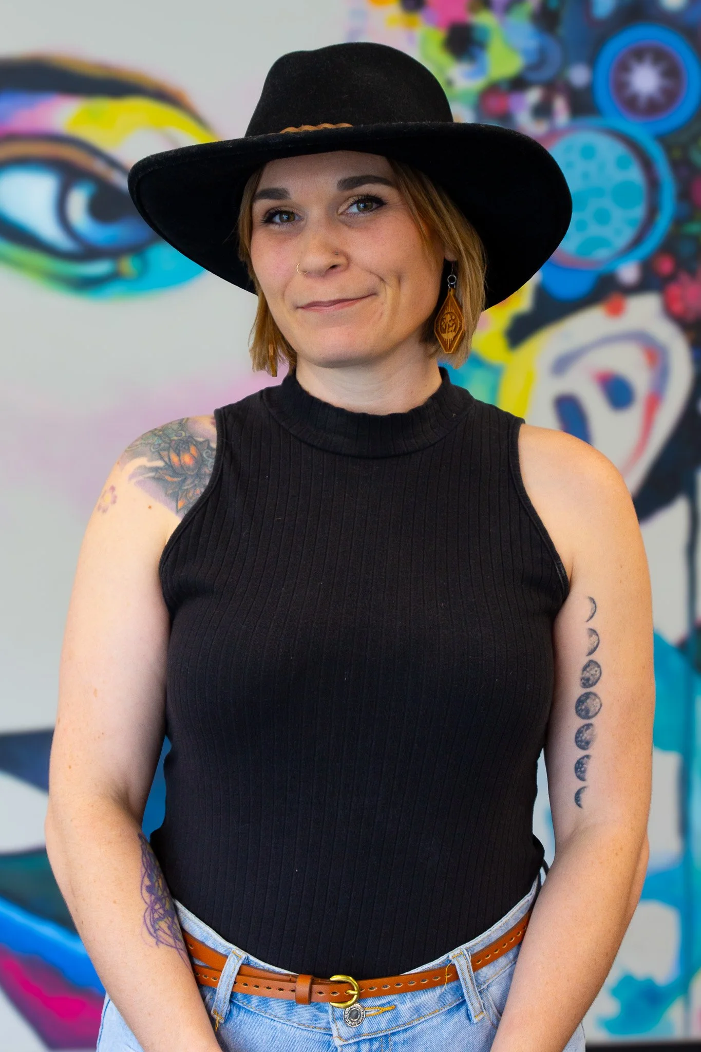 A woman with tattoos on her arm and wearing a black tank top, brown belt, and black wide-brimmed hat standing in front of a colorful abstract mural.
