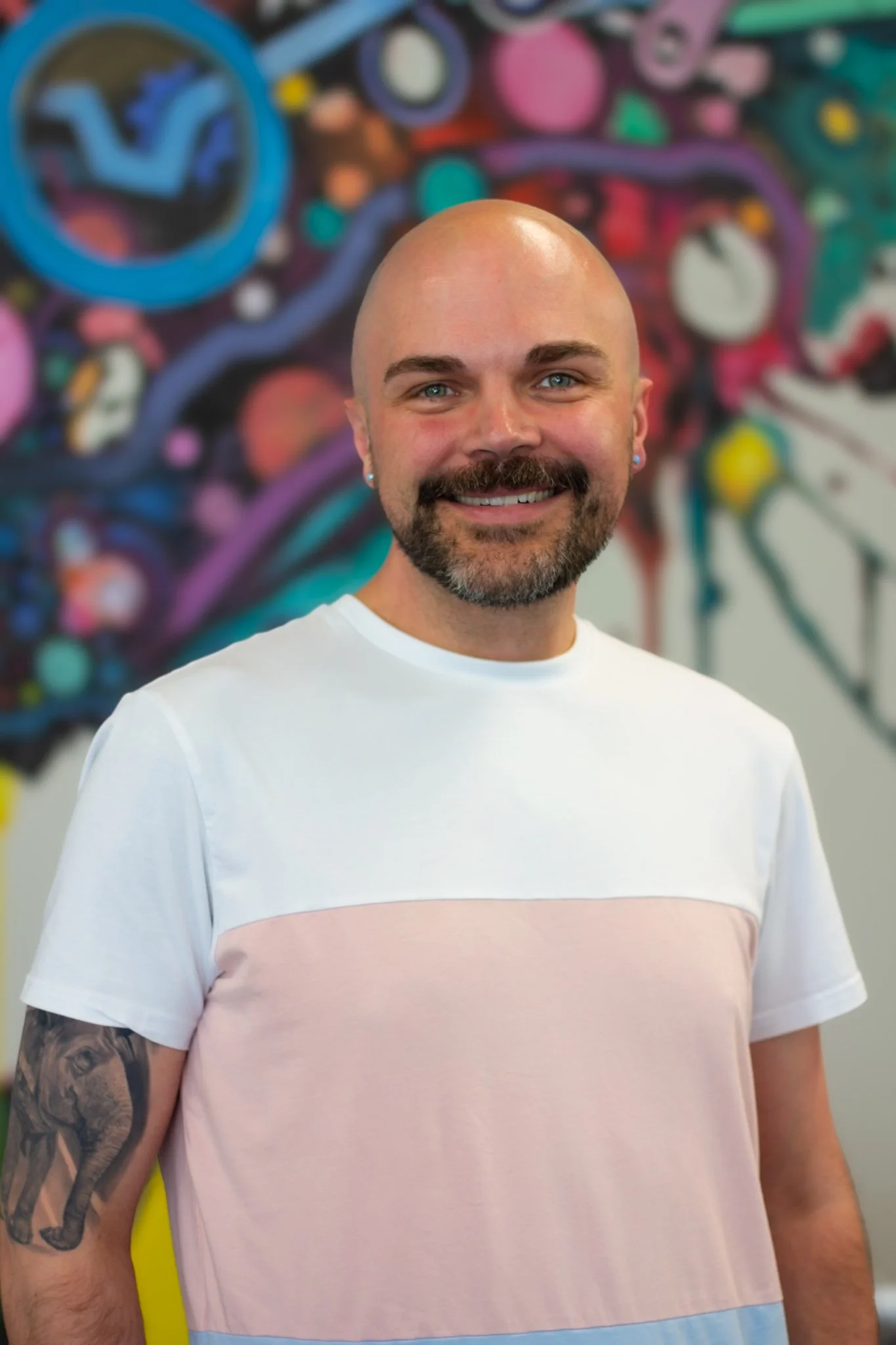 A smiling man with a shaved head and beard, wearing a white and pastel pink T-shirt, standing in front of a colorful graffiti wall.