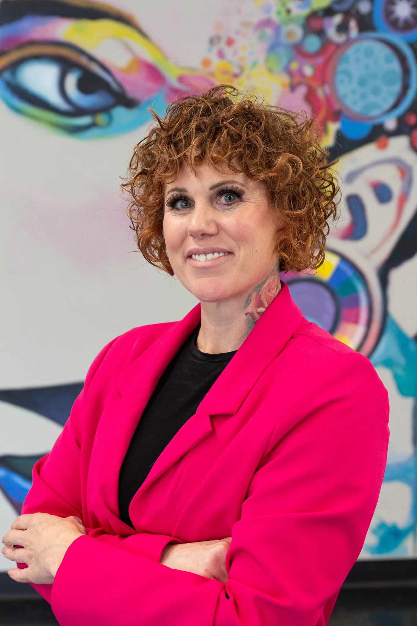A woman with curly red hair, wearing a pink blazer and black top, standing in front of a colorful abstract mural.