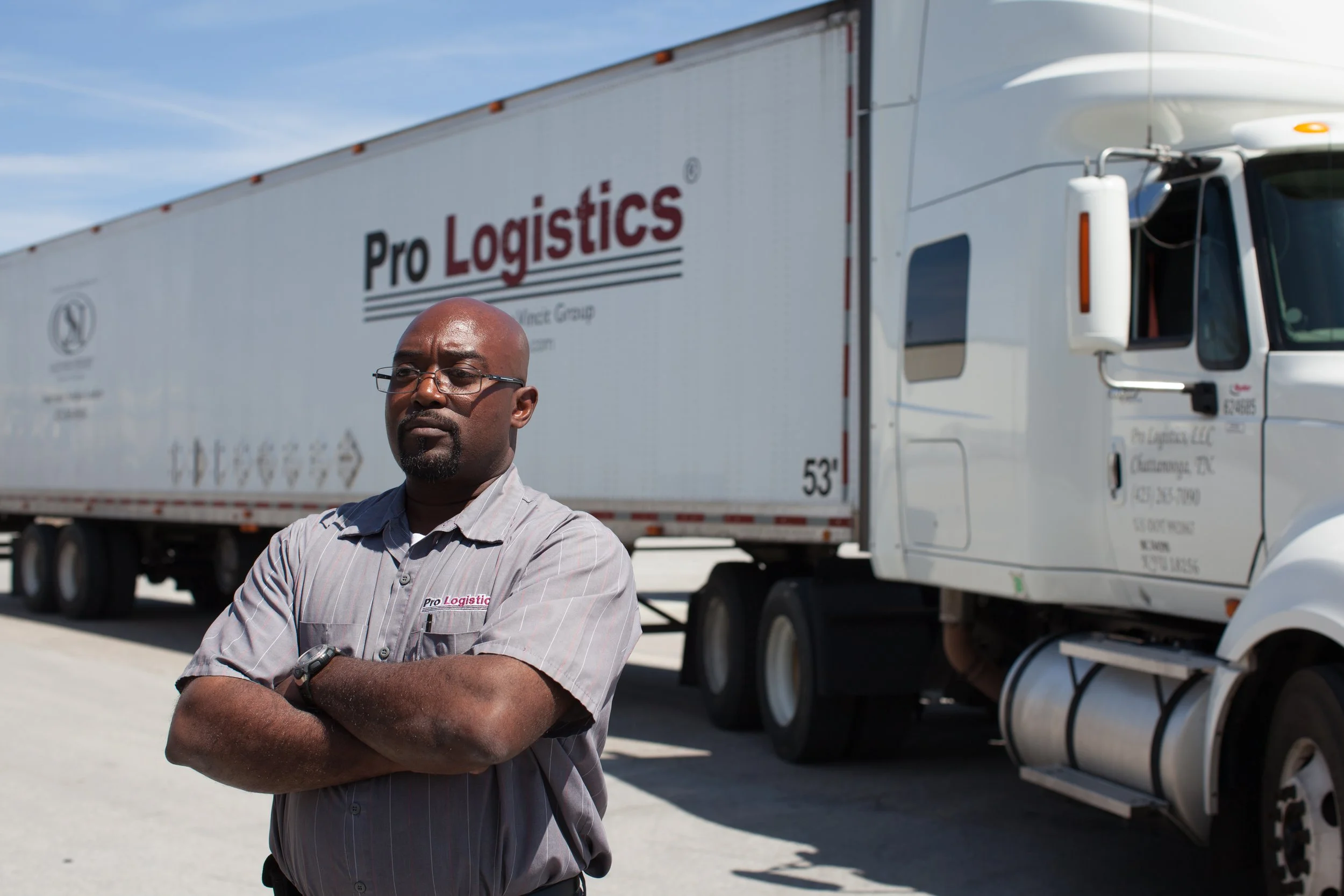A man with glasses and a gray striped shirt standing with arms crossed in front of a large white Pro Logistics truck on a sunny day.