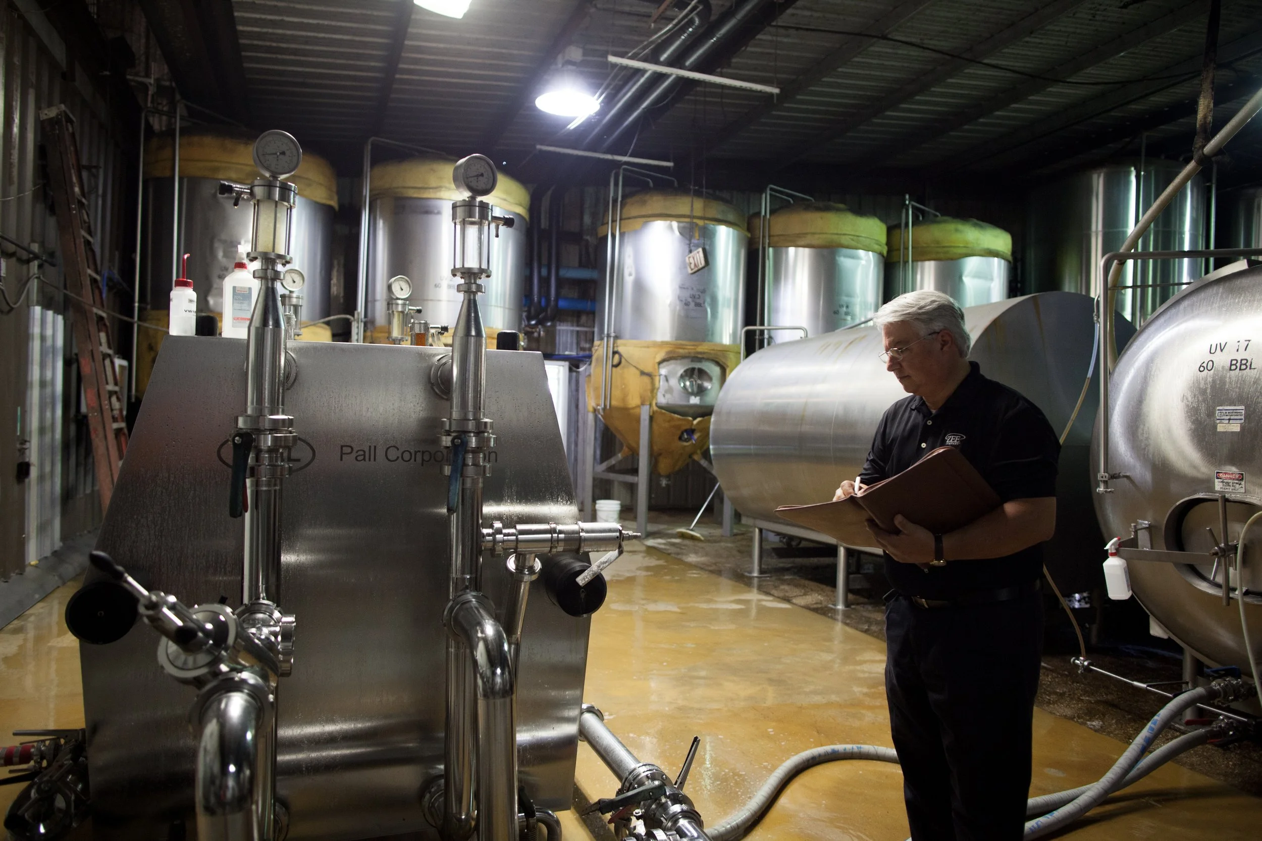 A man in a black polo shirt, reading a clipboard, stands in a brewery with large stainless steel fermentation tanks in the background.