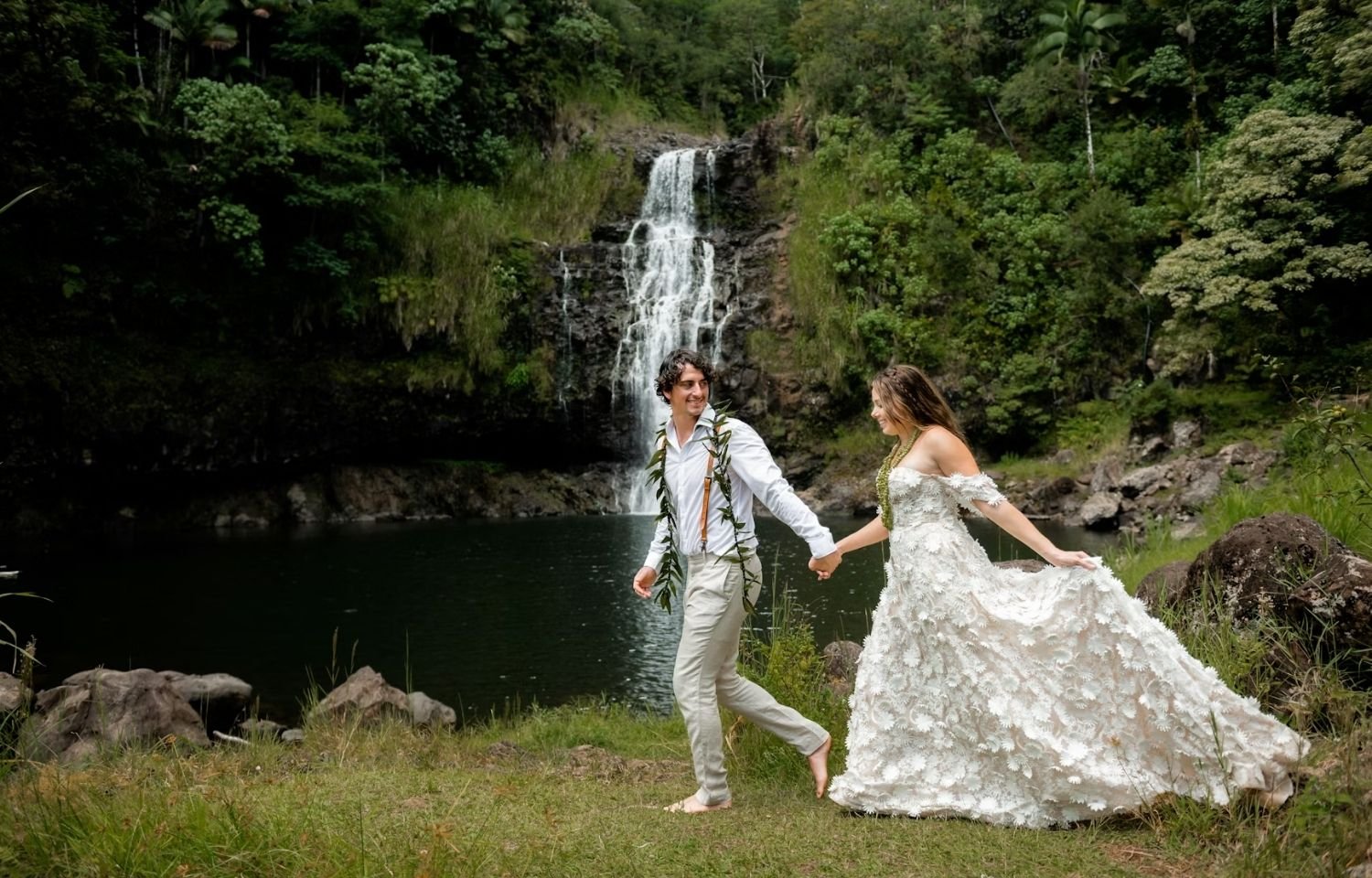 Bride and groom holding hands by tropical waterfall, outdoor nature-inspired Hawaii wedding. Unique elopement ideas for couples seeking scenic ceremony locations.