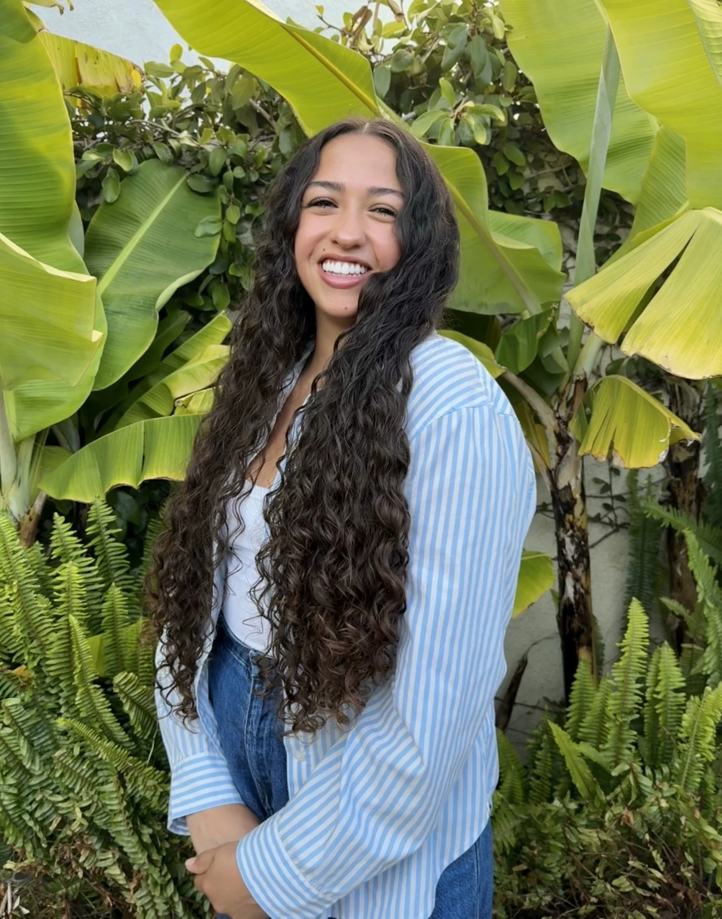 A young woman with long, curly dark hair smiling outdoors among large green banana leaves and ferns, wearing a light blue striped shirt and blue jeans.
