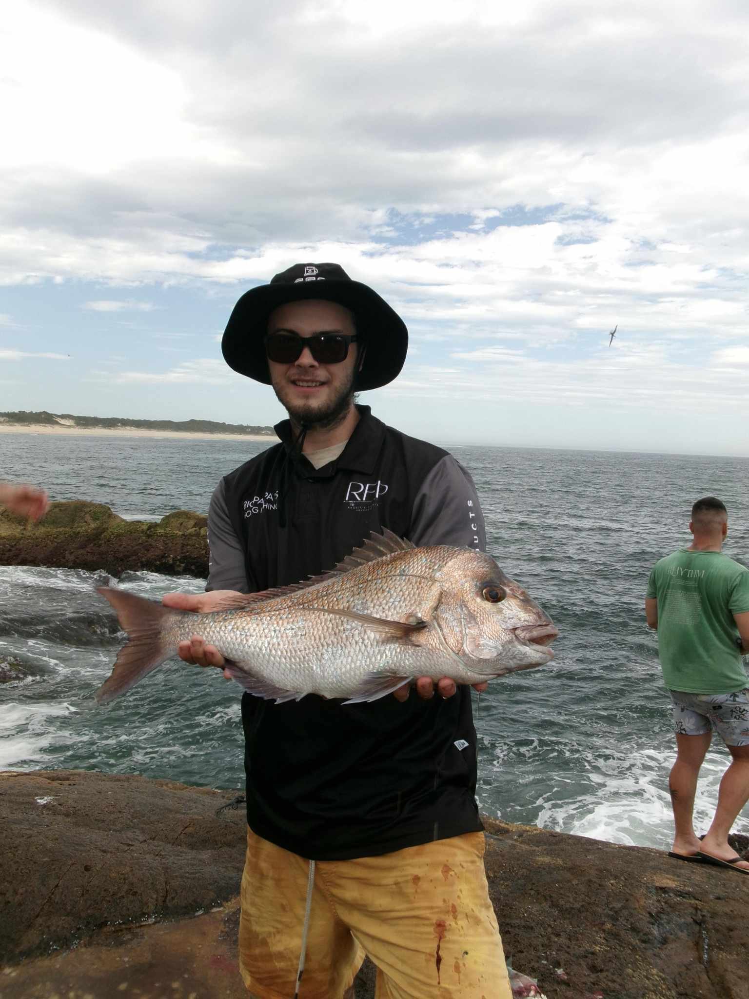 A man wearing sunglasses, a black sunhat, and a black jacket is holding a large fish near the rocky shoreline of the ocean. Other people are visible in the background, with one person in a green shirt and shorts standing on the rocks, and seagulls flying overhead with a cloudy sky above.