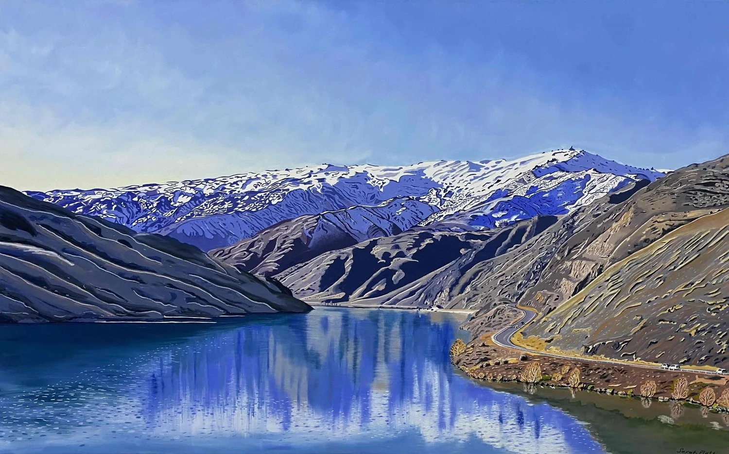 Scenic landscape of snow-capped mountains, a winding road, and a calm river reflecting the surrounding hills under a clear blue sky.