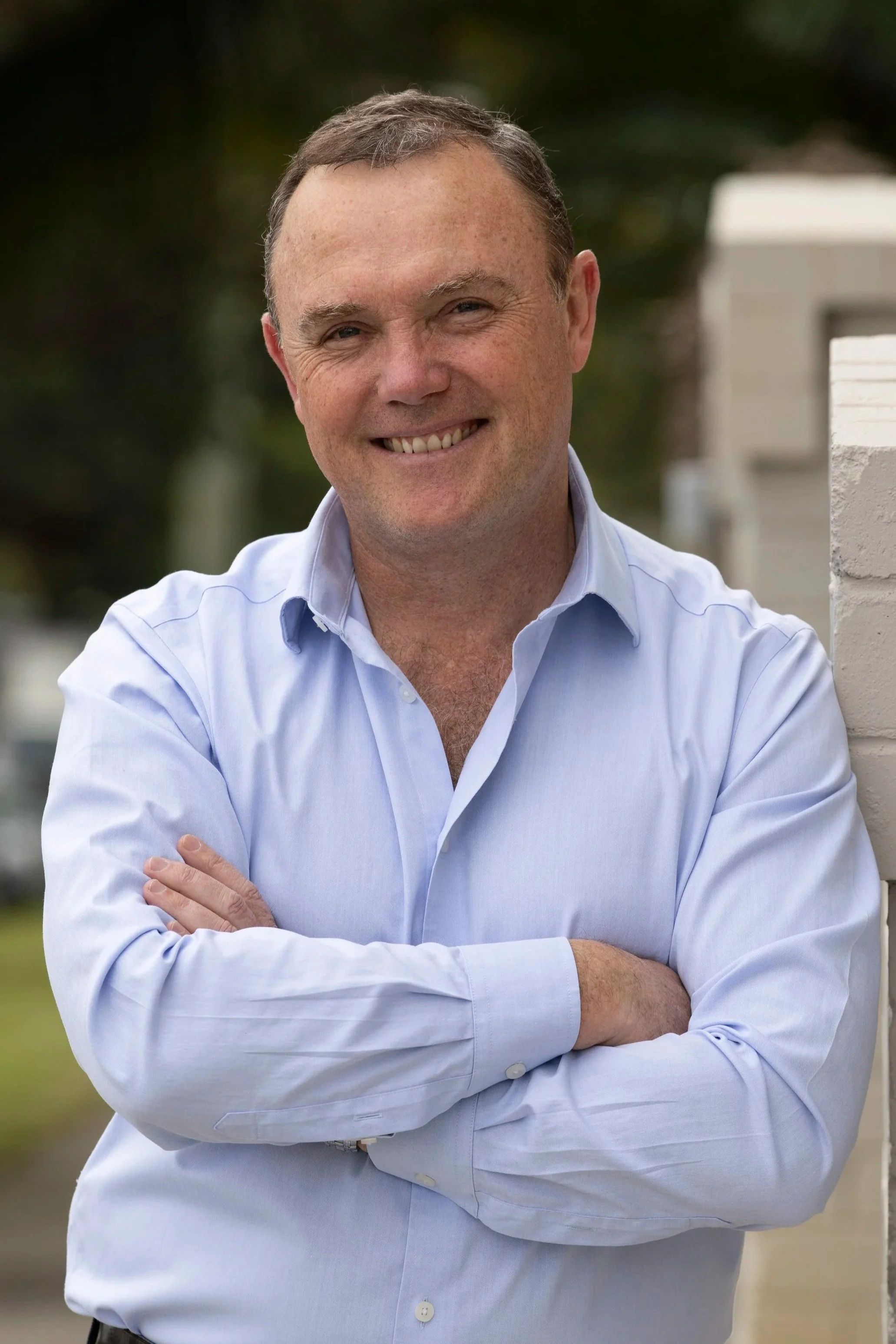Smiling man in a blue checkered shirt standing in front of green foliage.