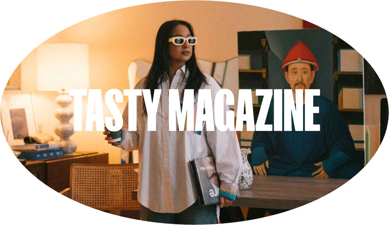 Woman with long dark hair and glasses holding a coffee and a magazine, standing in a warmly lit living room with a lamp, picture frame, and books. In the background, a man in a blue outfit and red hat with elf-like ears is sitting at a table with a green candle and a TV.