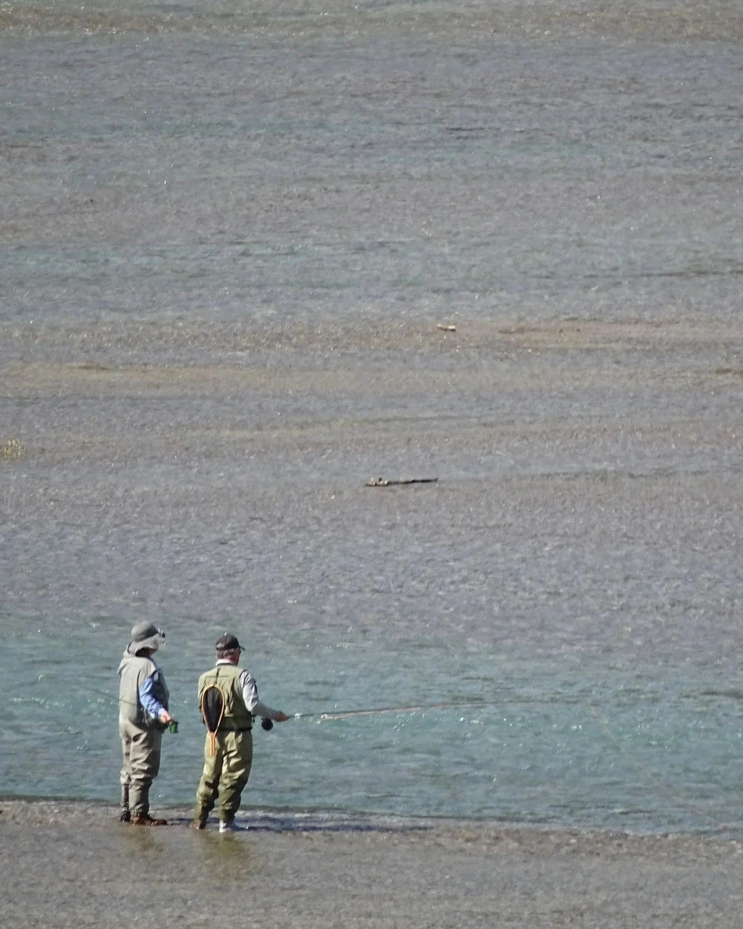 Two people in fishing gear standing by the water on a rocky beach, with a vast expanse of water and shoreline in the background.
