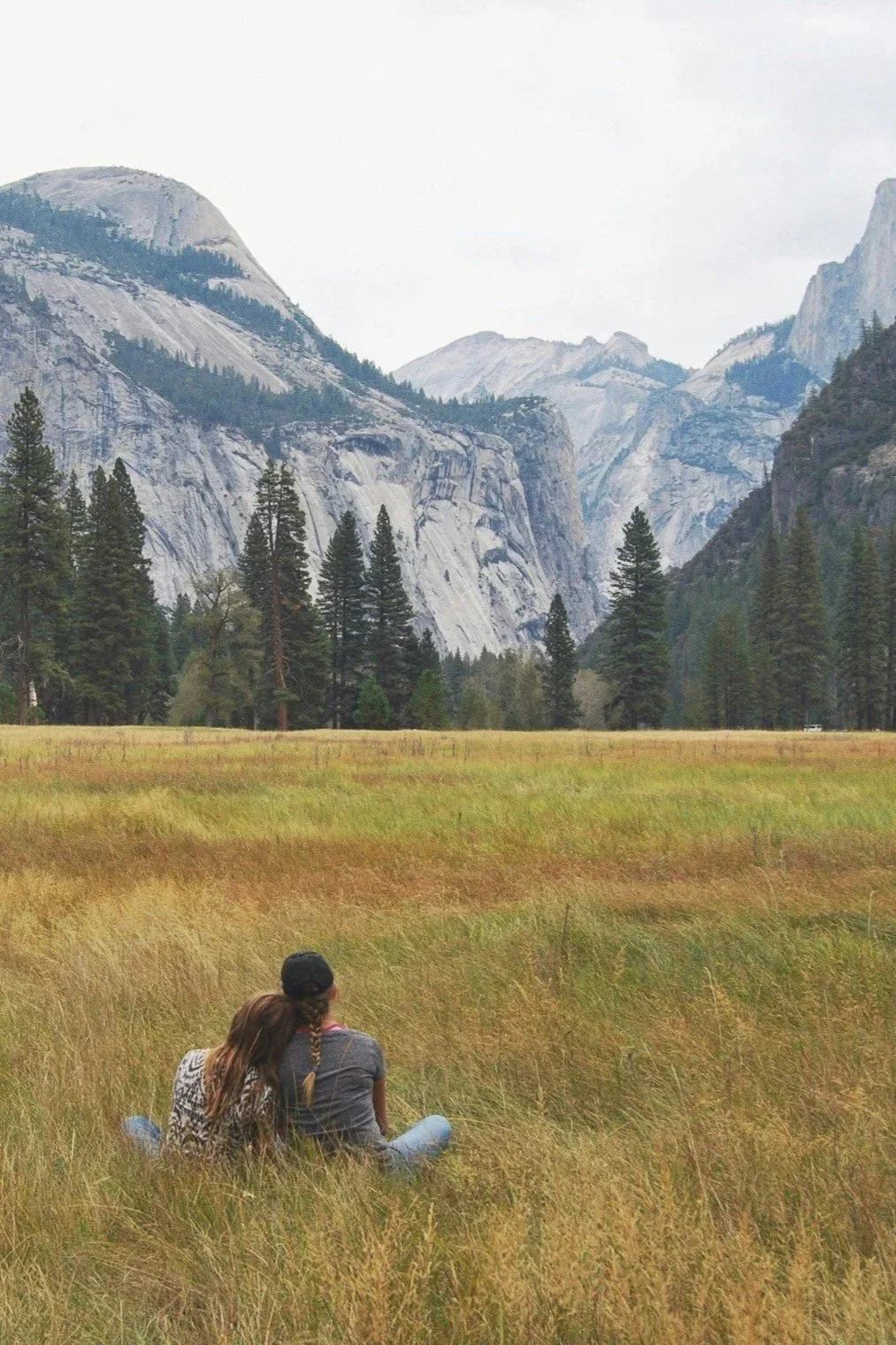 Two people sitting in a grassy field with a backdrop of trees, mountains, and a cloudy sky.