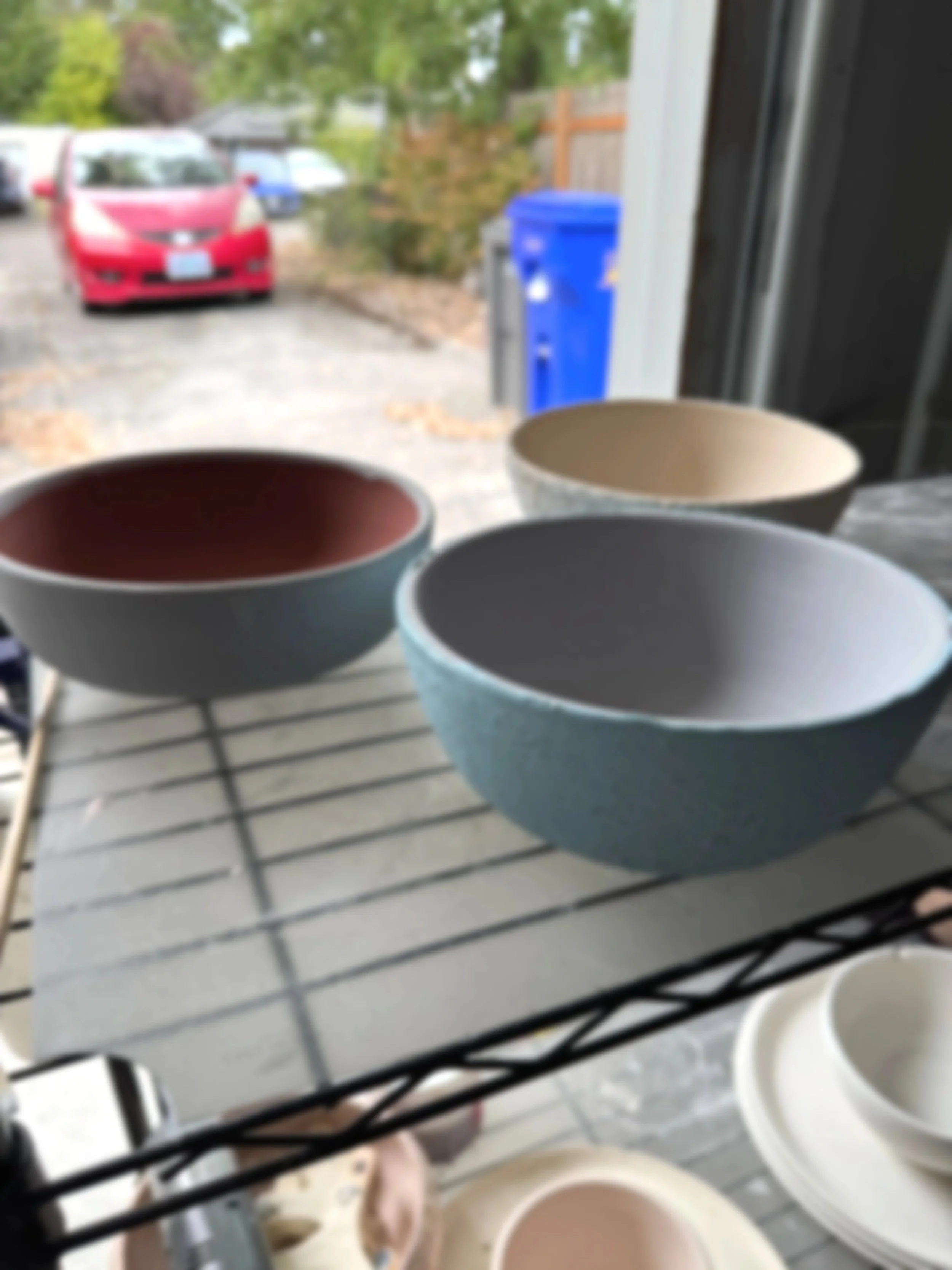 Colorful ceramic bowls on a wire shelf in a garage with a view of parked cars and trees outside.