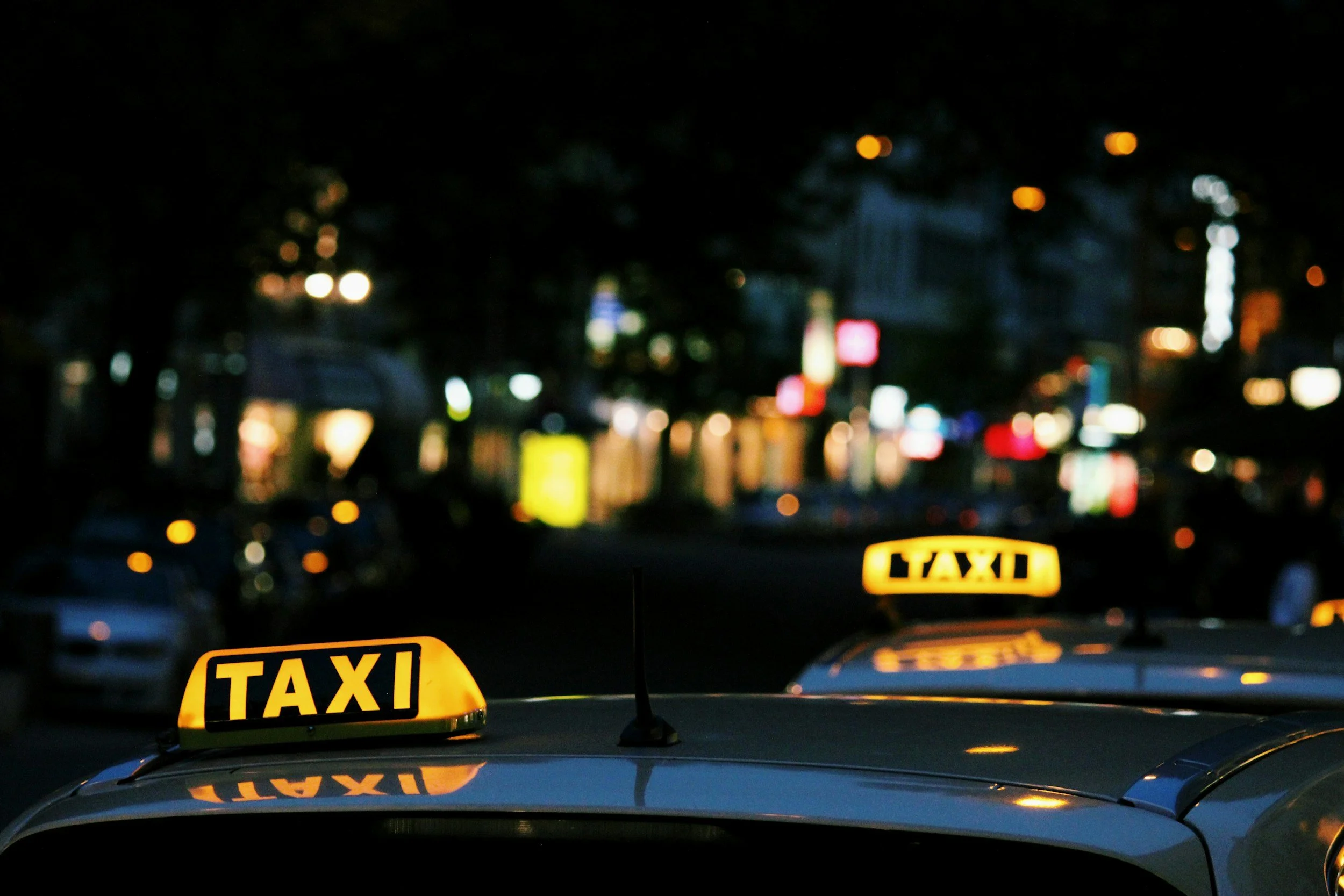 Night scene of two illuminated yellow taxi signs on the roofs of parked taxis on a city street.