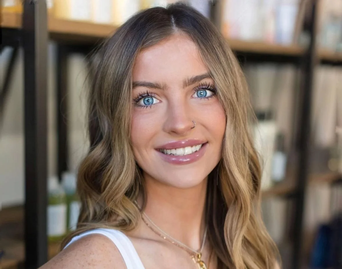 A woman with blue eyes, light brown hair, and a nose piercing smiling at the camera, with a bookshelf in the background.