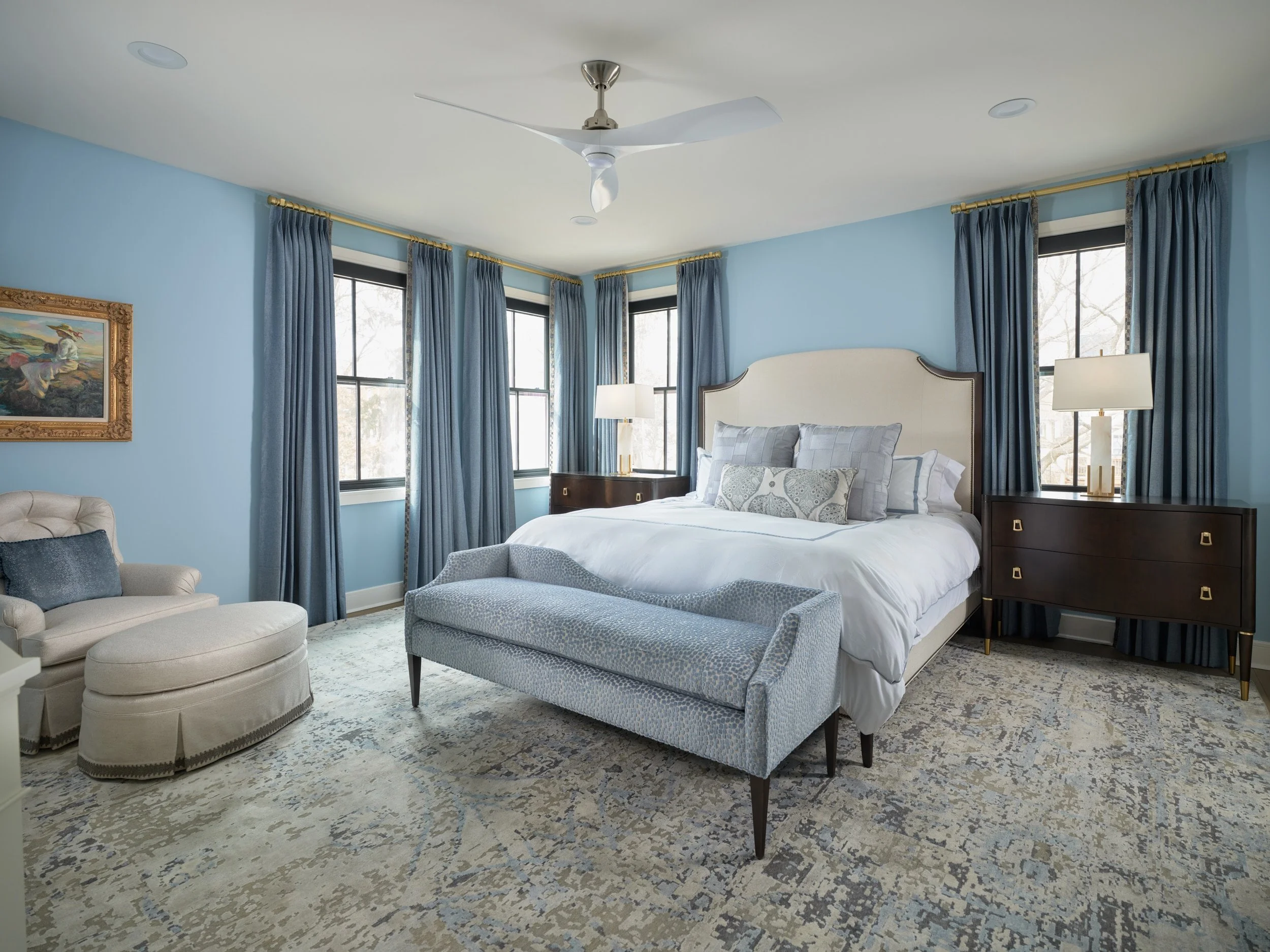 A bedroom with light blue walls, four tall windows with blue curtains, and a white bed with multiple patterned pillows. There is a beige upholstered chair, a dark wood nightstand with a lamp, and a dresser. A ceiling fan and framed painting are also visible.
