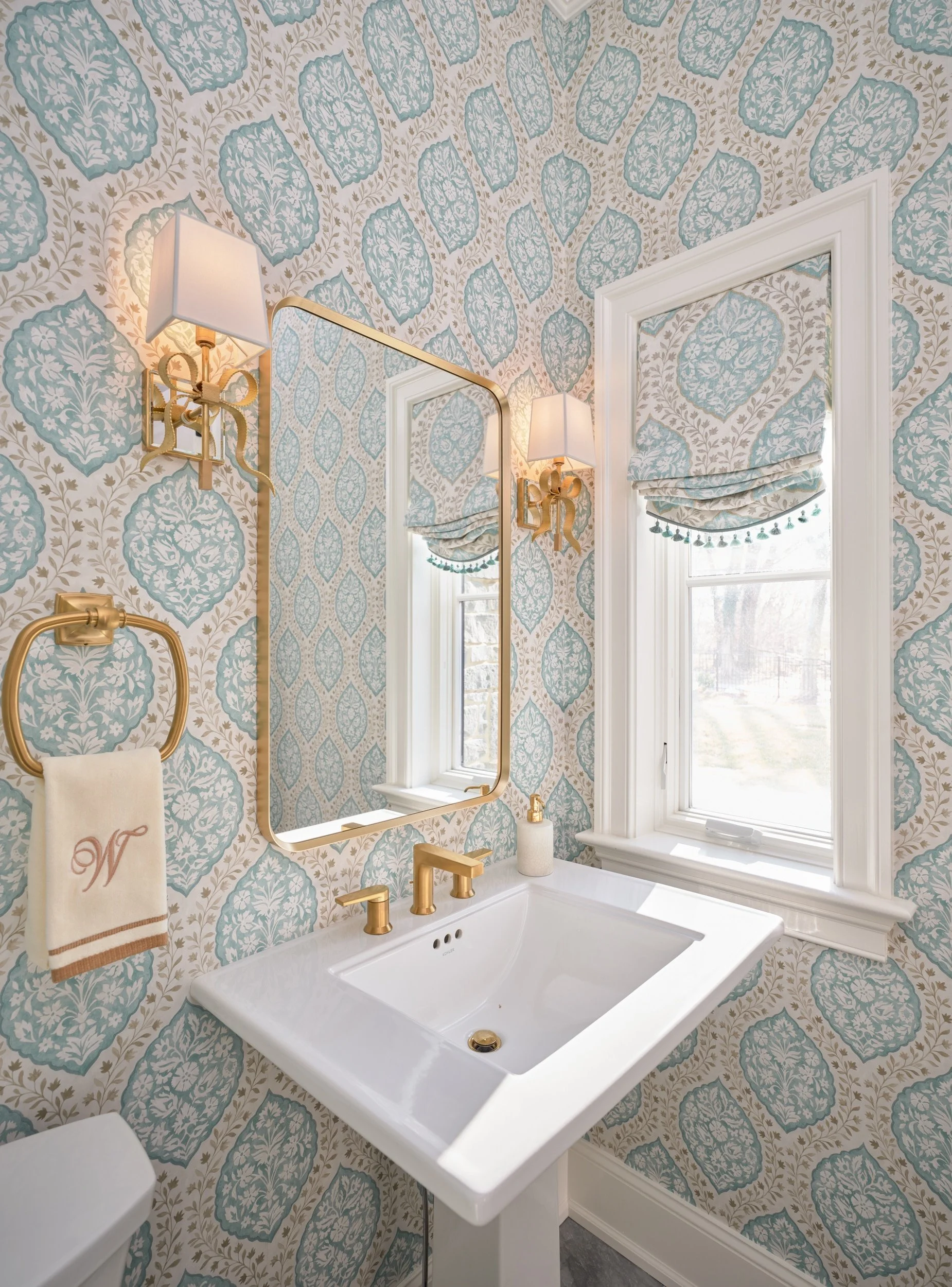 A bathroom with patterned wallpaper featuring blue and beige floral motifs, a white rectangular sink, a large window with a Roman shade, and gold fixtures including two wall-mounted lamps, a faucet, and decorative towel rings.