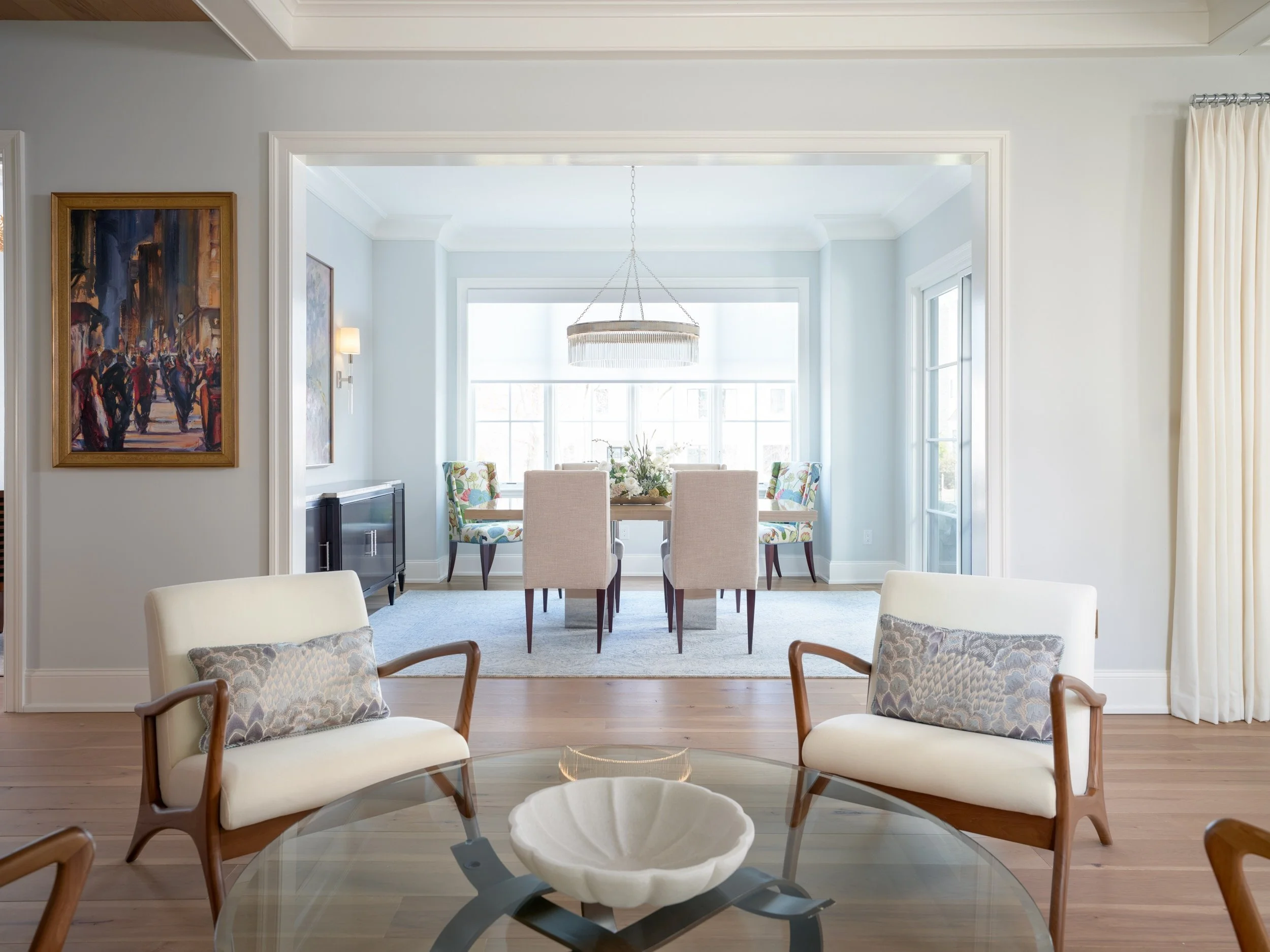 A view of a dining area with a table, chairs, and a chandelier. In the foreground, there are two beige chairs with patterned pillows. The room has large windows letting in natural light and a light-colored wall with artwork on the left.
