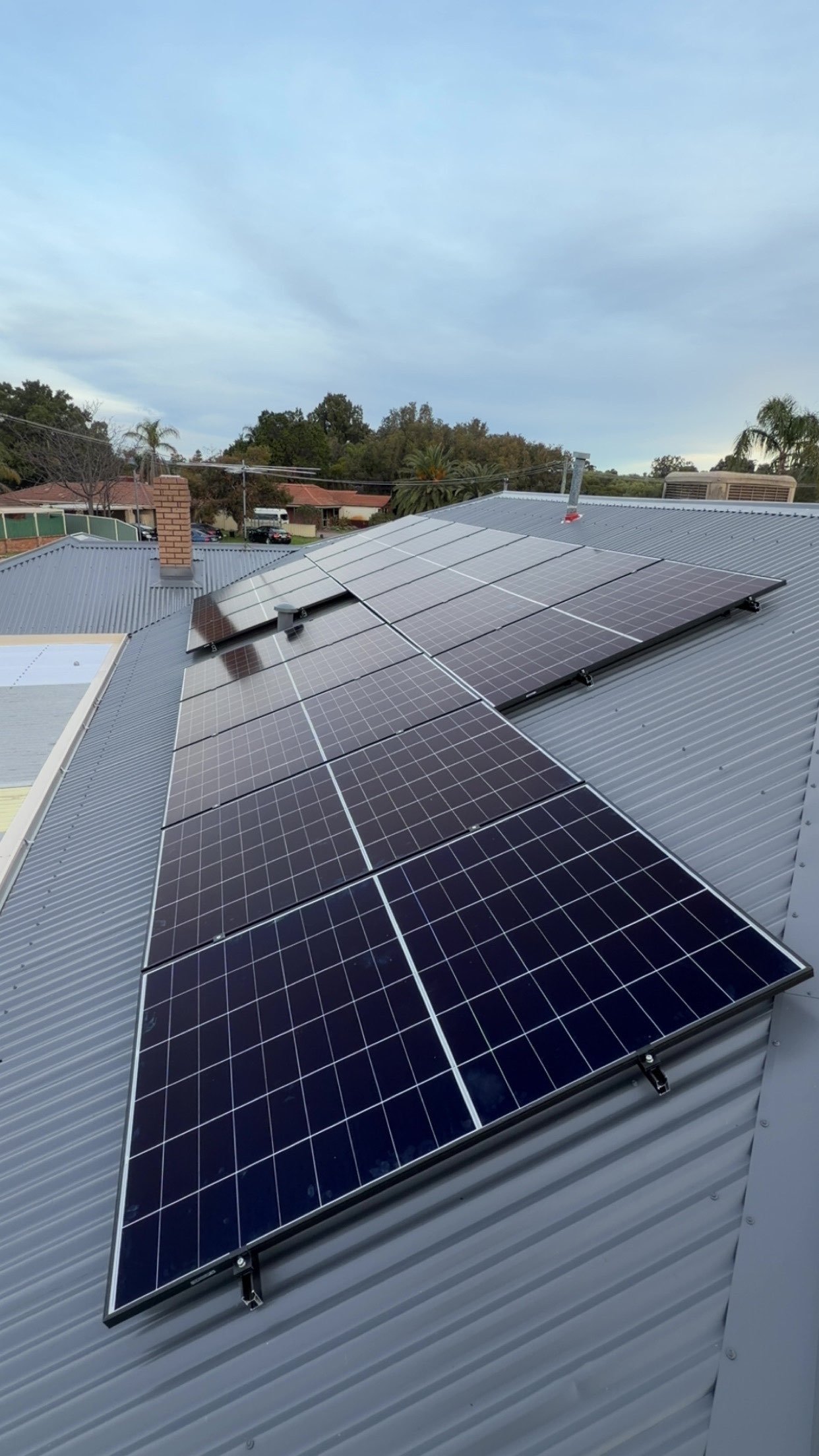 Solar panels installed on a metal roof with a background of houses, trees, and a cloudy sky.