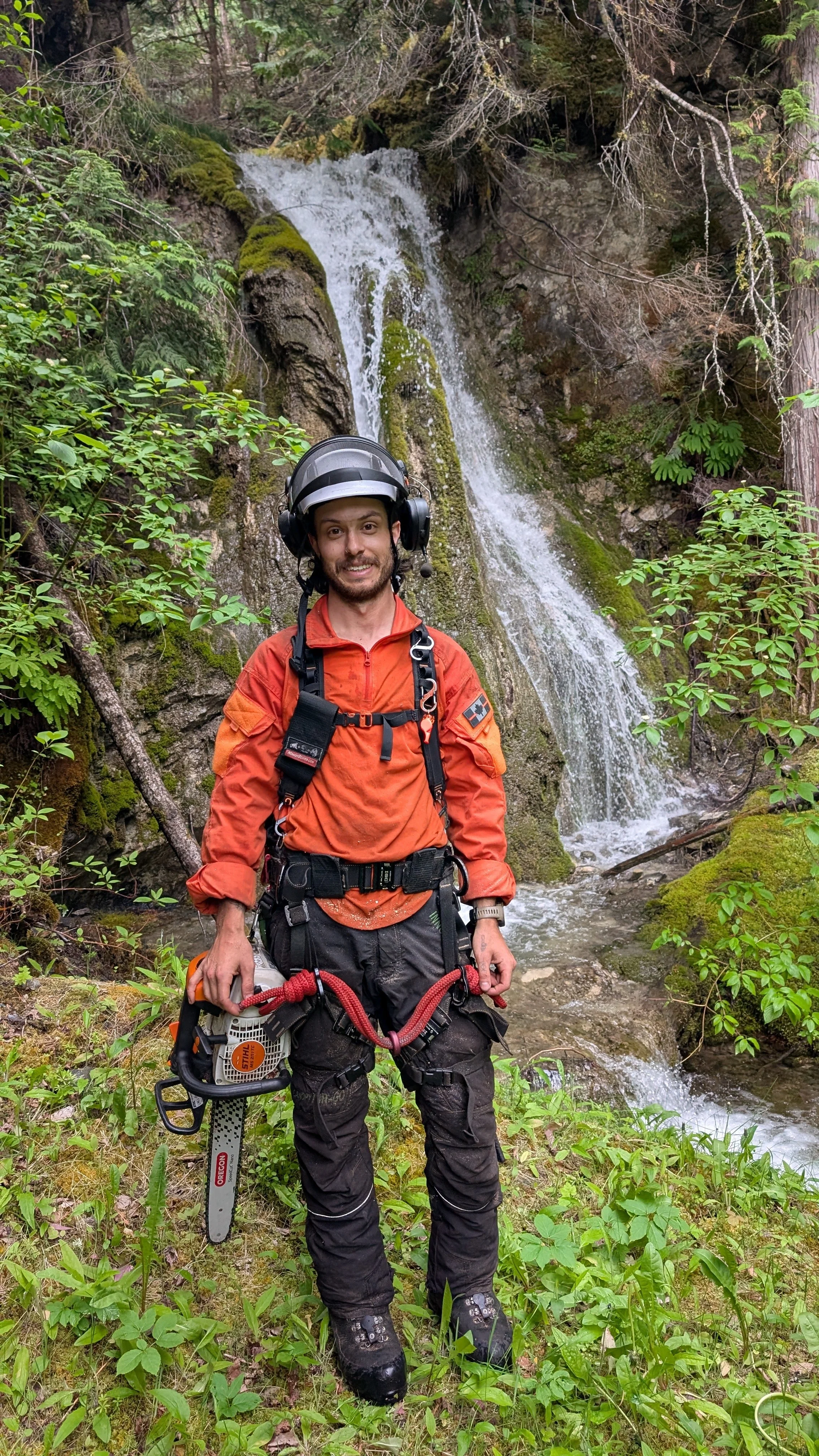 A man in outdoor gear, holding a chainsaw, standing in front of a waterfall surrounded by greenery in a forest.