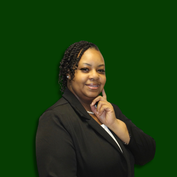 A woman with twisted hair, wearing a black blazer and pearl necklace, standing against a dark green background with a confident smile.