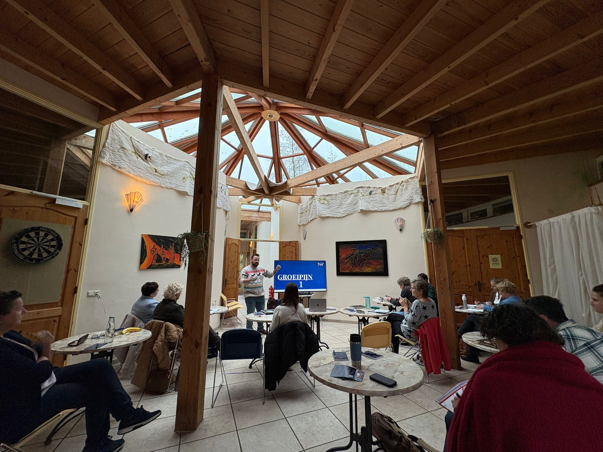 Binnenruimte met houten plafond en een glazen koepel, mensen zitten aan ronde tafels, een man geeft een presentatie voor een scherm met tekst, decoratieve lampen en muurkunst