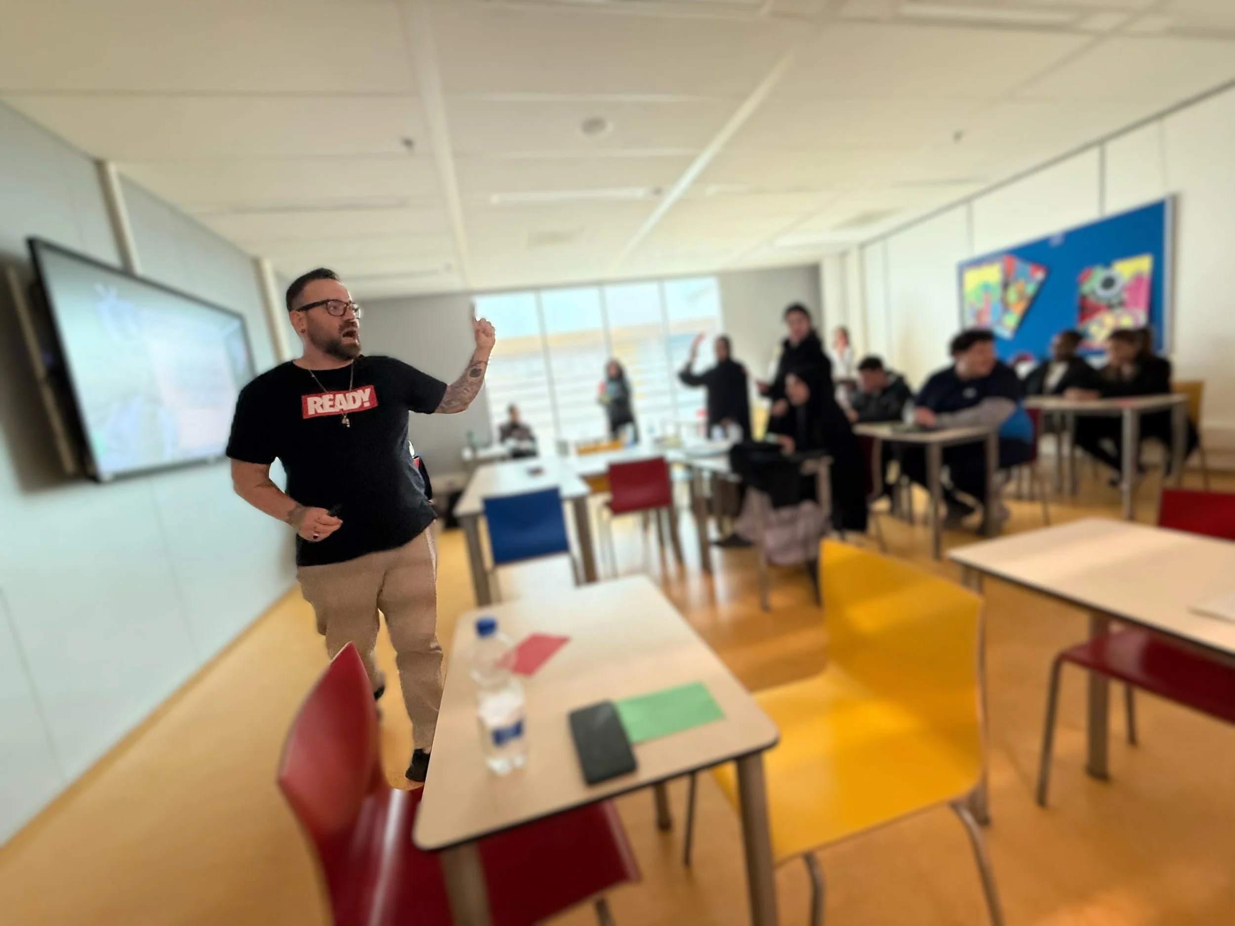 Man geeft les aan een groep studenten in een klaslokaal, met een whiteboard en kleurrijke kunstwerken aan de muur